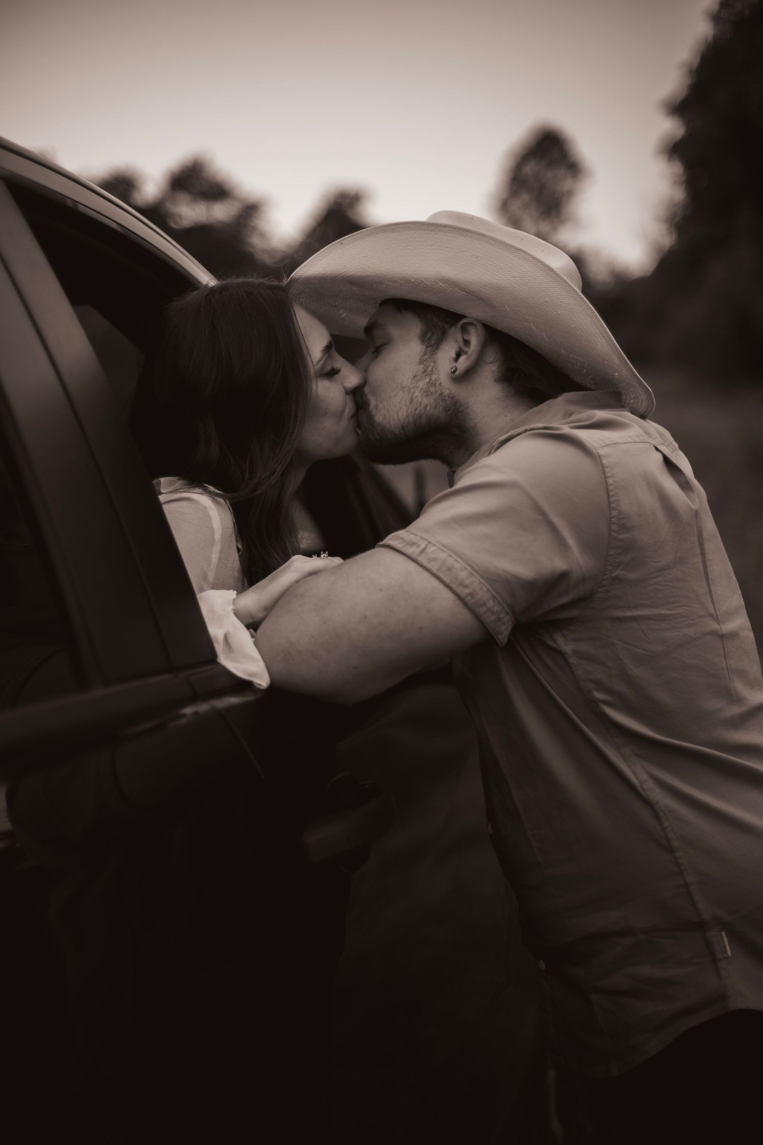Sepia-toned photo of a couple sharing a romantic kiss beside a truck, man wearing a cowboy hat outdoors.
