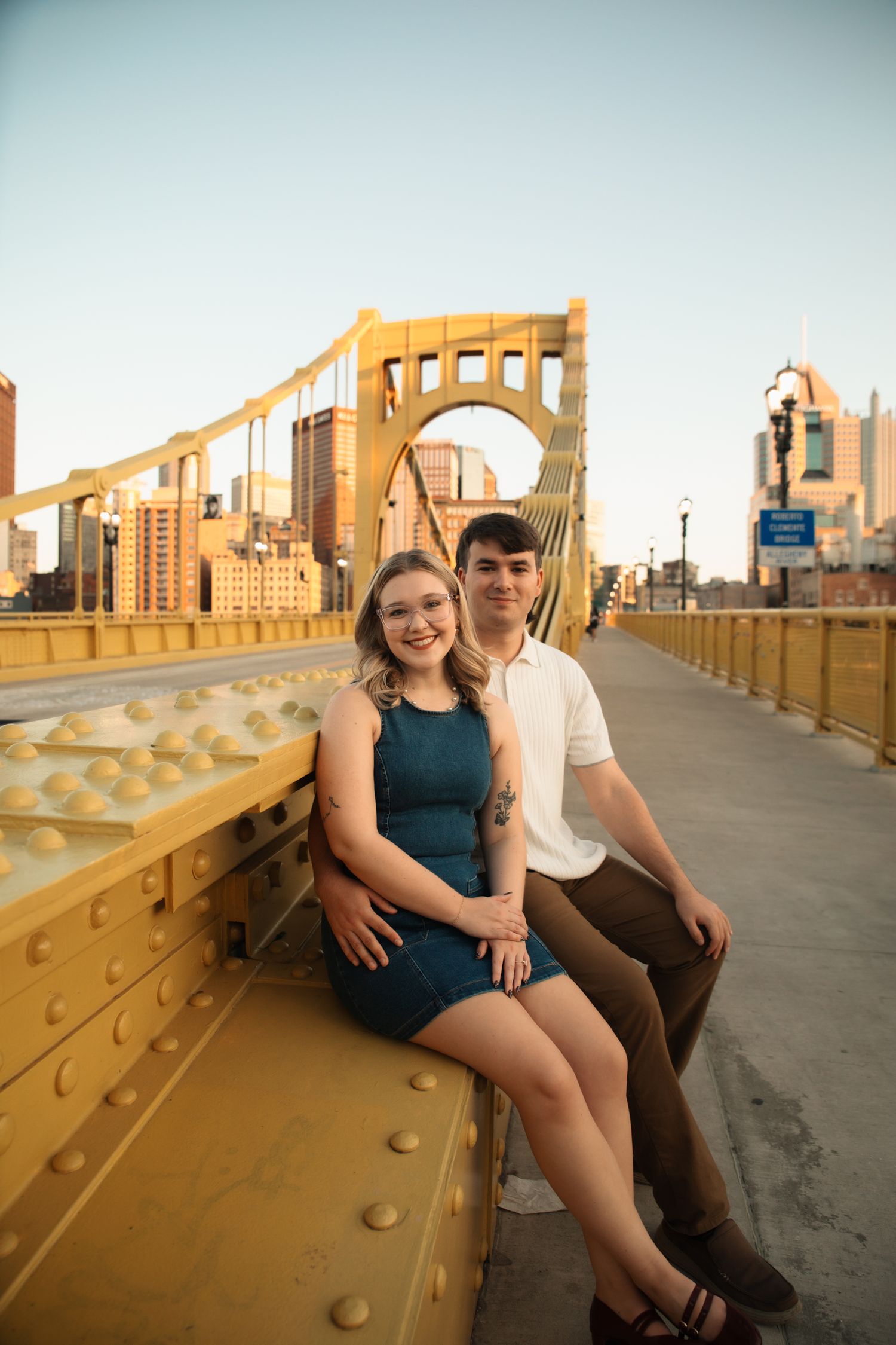 A couple sits together on Pittsburgh's iconic yellow Roberto Clemente Bridge at sunset with the city skyline behind them.