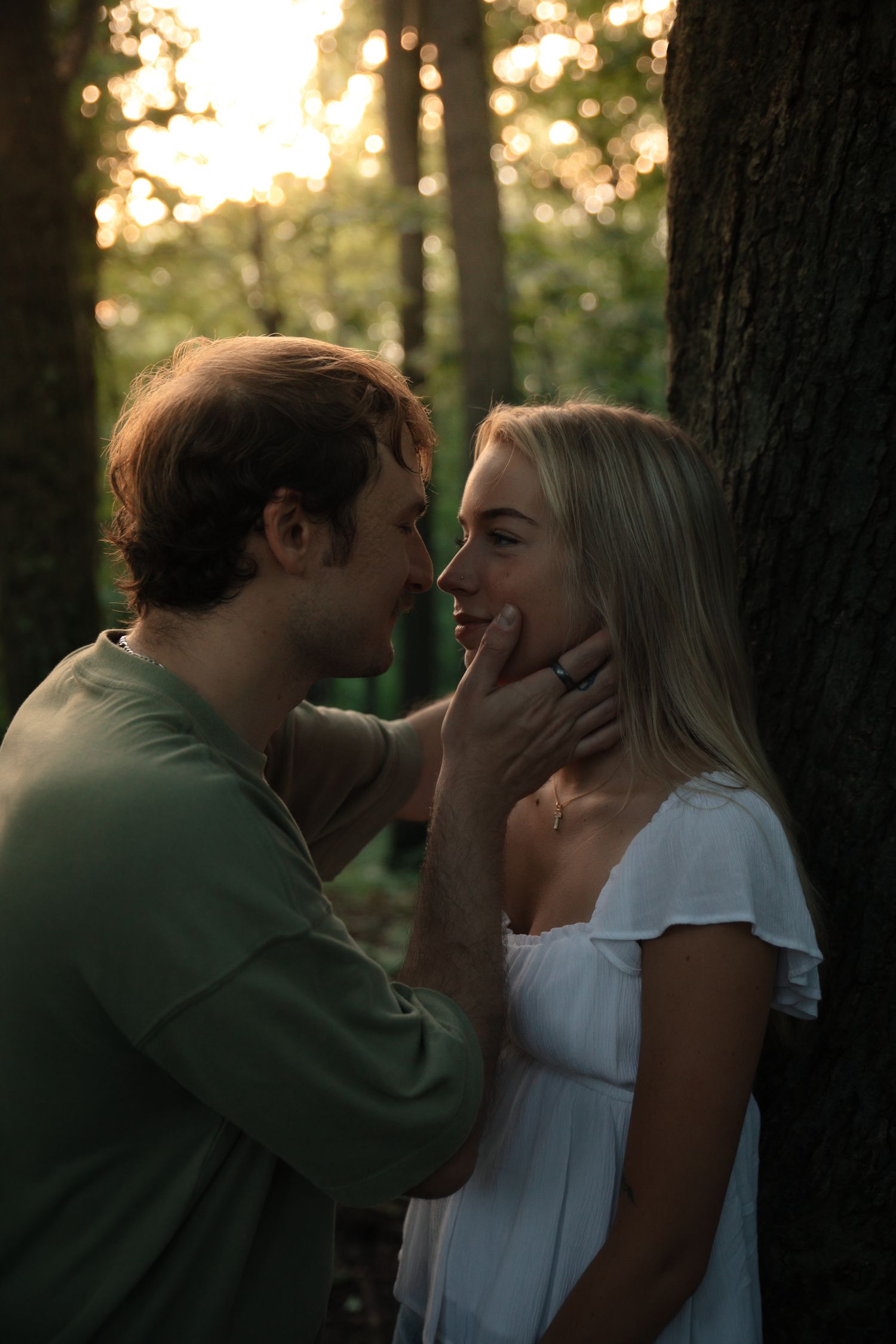 A romantic couple shares an intimate moment in a sunlit forest with bokeh lighting effects in the background.