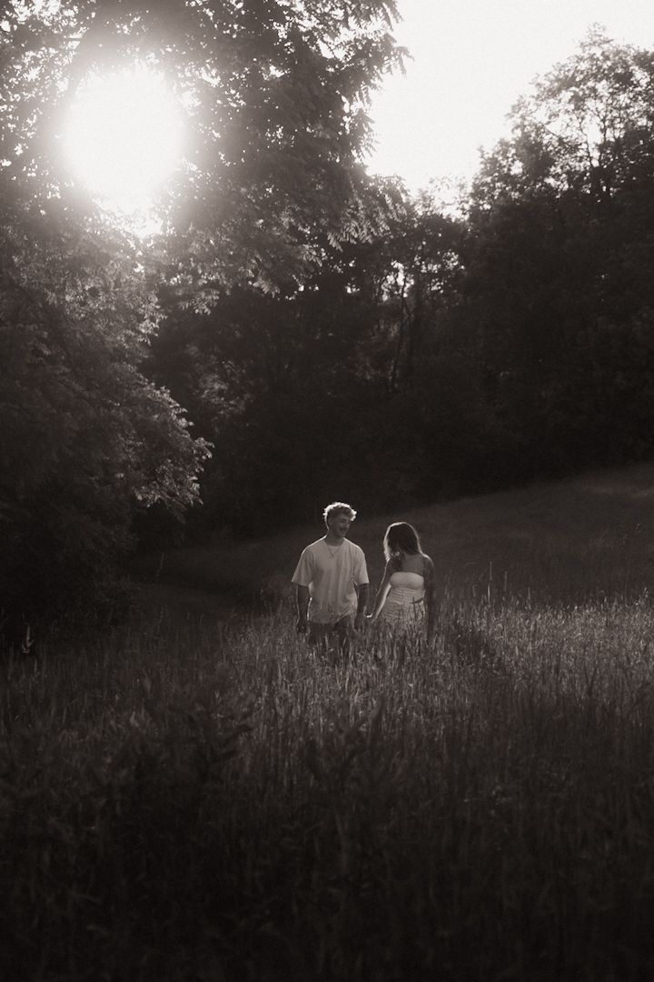 Black and white photo of a couple holding hands walking through a sunlit meadow surrounded by lush trees.