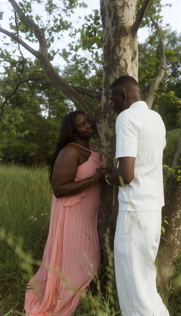 A couple in a park, woman in pink dress and man in white outfit, holding hands by a tree.