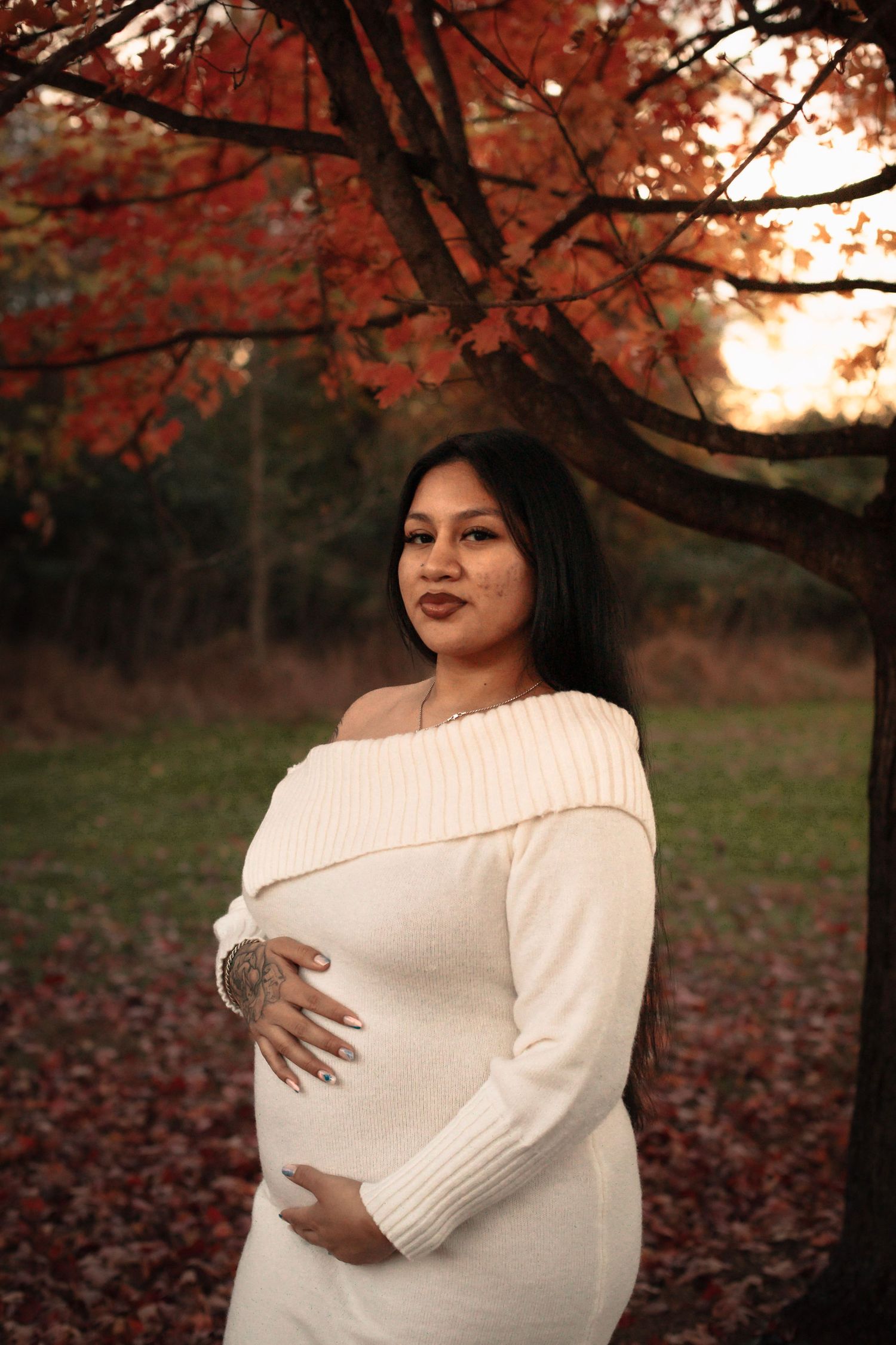 A person in a white sweater poses outdoors under autumn trees with red foliage at sunset.