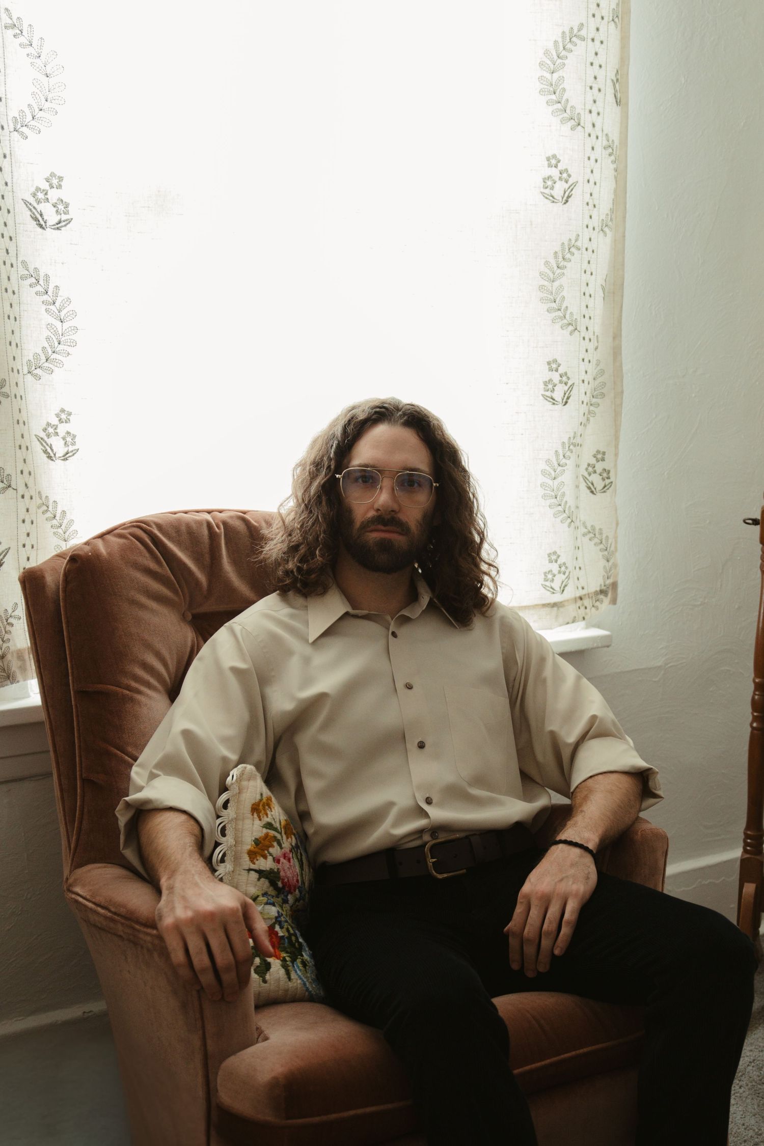 Man with long curly hair and glasses sitting in a vintage armchair near a curtained window wearing a beige shirt.