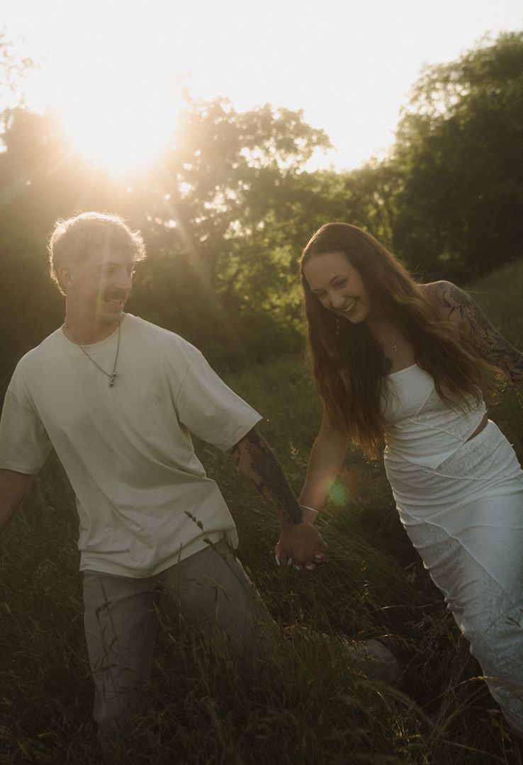 A couple holds hands walking through a sunlit field at golden hour, she wears white dress, he wears cream tee.