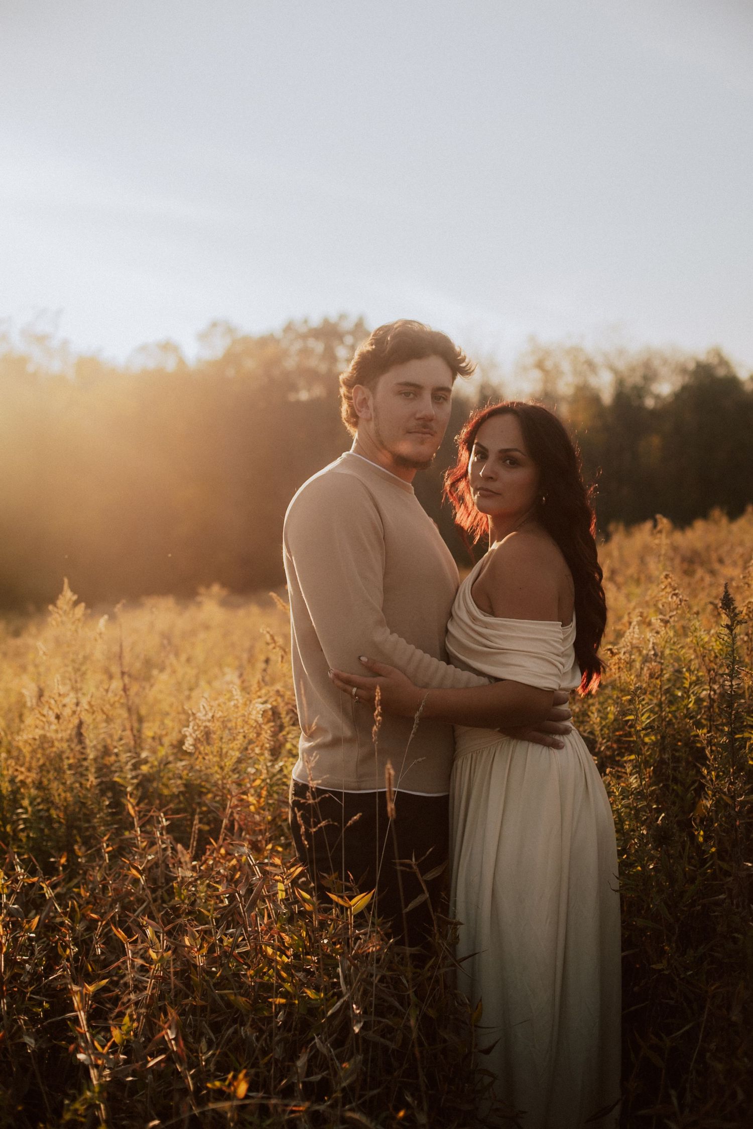 A couple embraces in a golden field at sunset with warm backlight illuminating the autumn wildflowers.