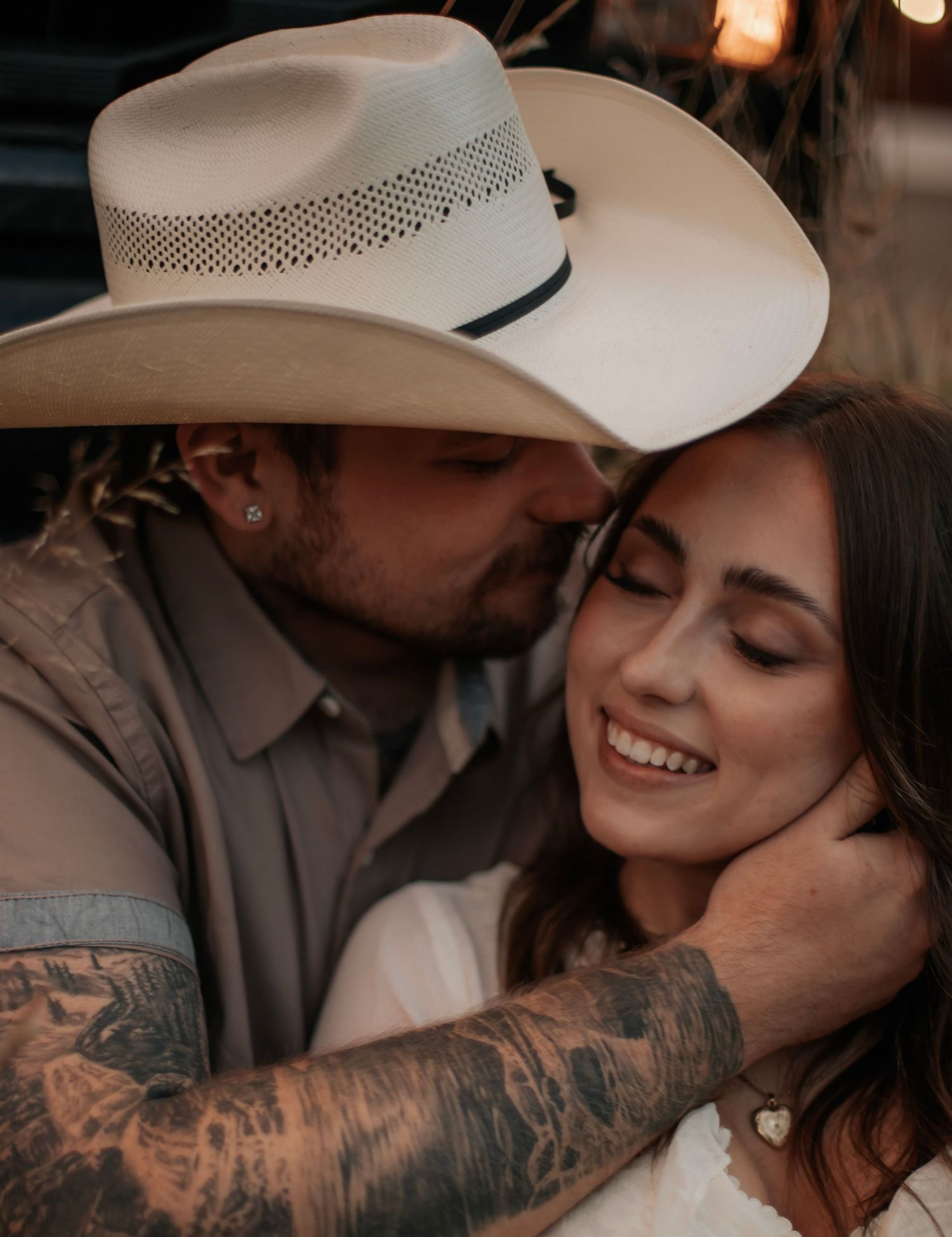 A tattooed man in a cowboy hat kisses a smiling woman tenderly on the cheek in a romantic outdoor portrait.