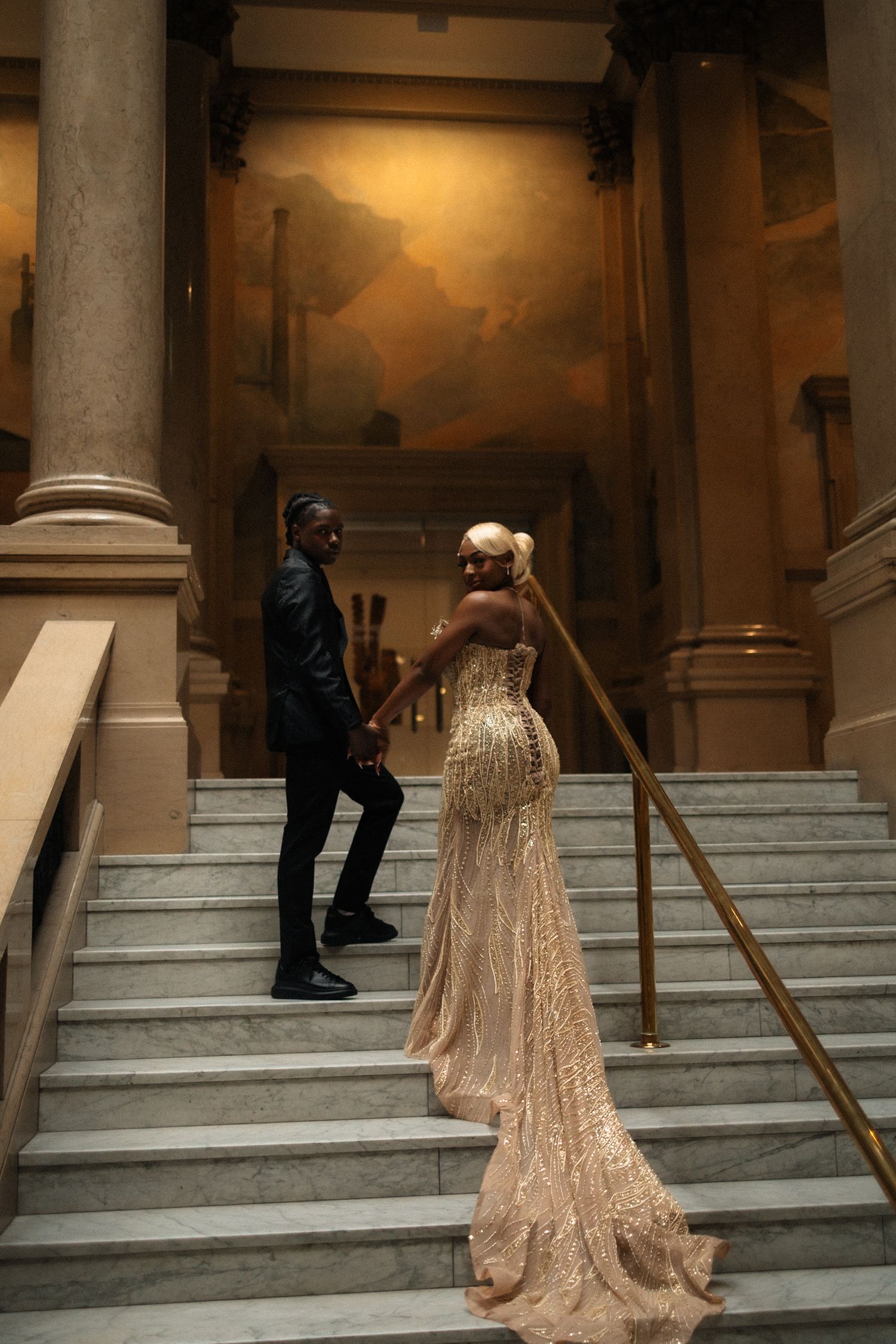 Elegant couple in formal attire holding hands on marble stairs, woman in gold sequin gown with train, grand hall backdrop.
