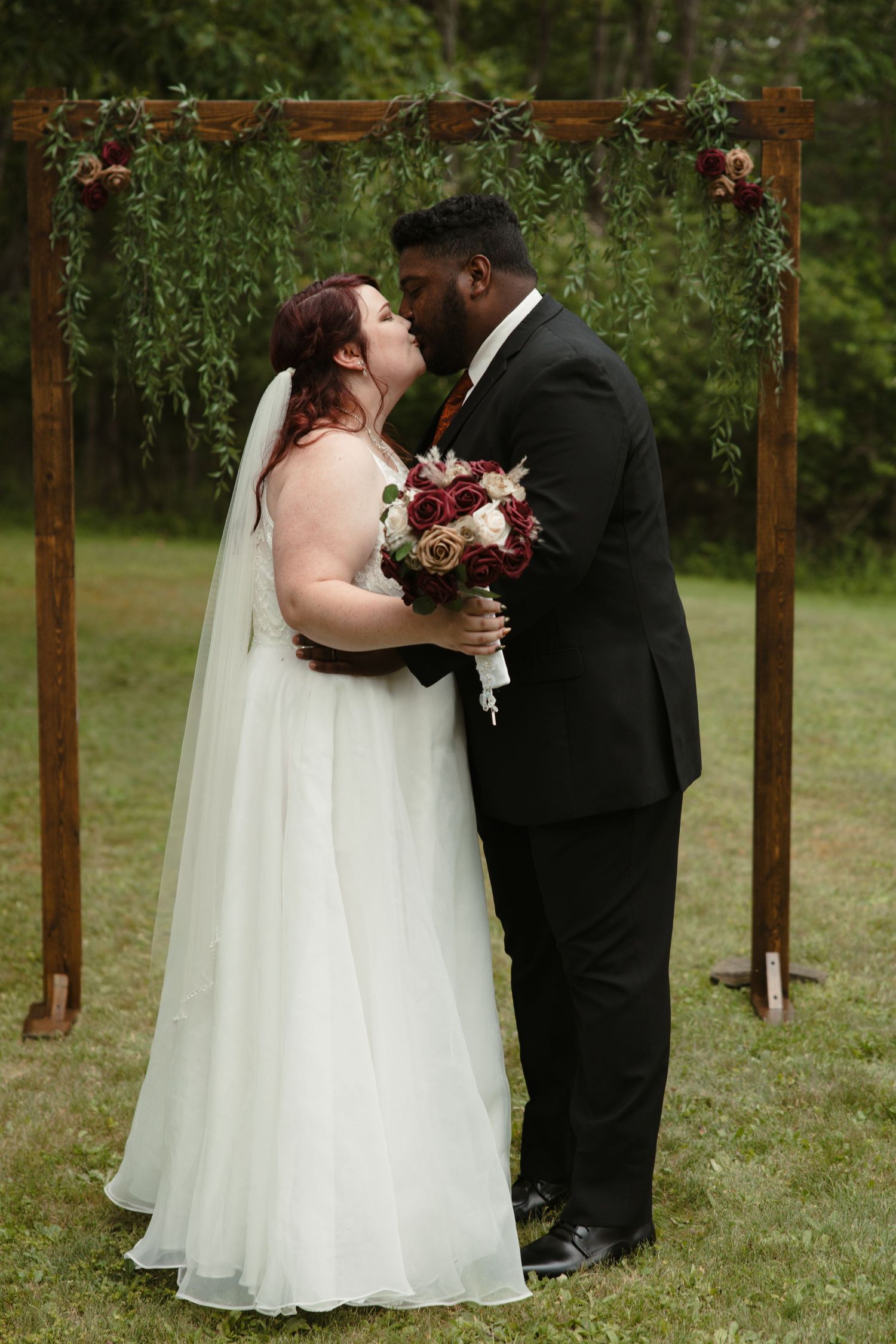 A bride and groom share their first kiss under a rustic wooden arch adorned with greenery and burgundy flowers at an outdoor wedding.
