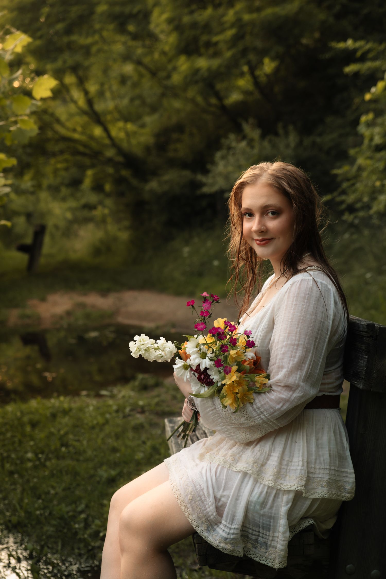 Young woman in white dress holding colorful wildflower bouquet, seated on bench in lush green park setting.