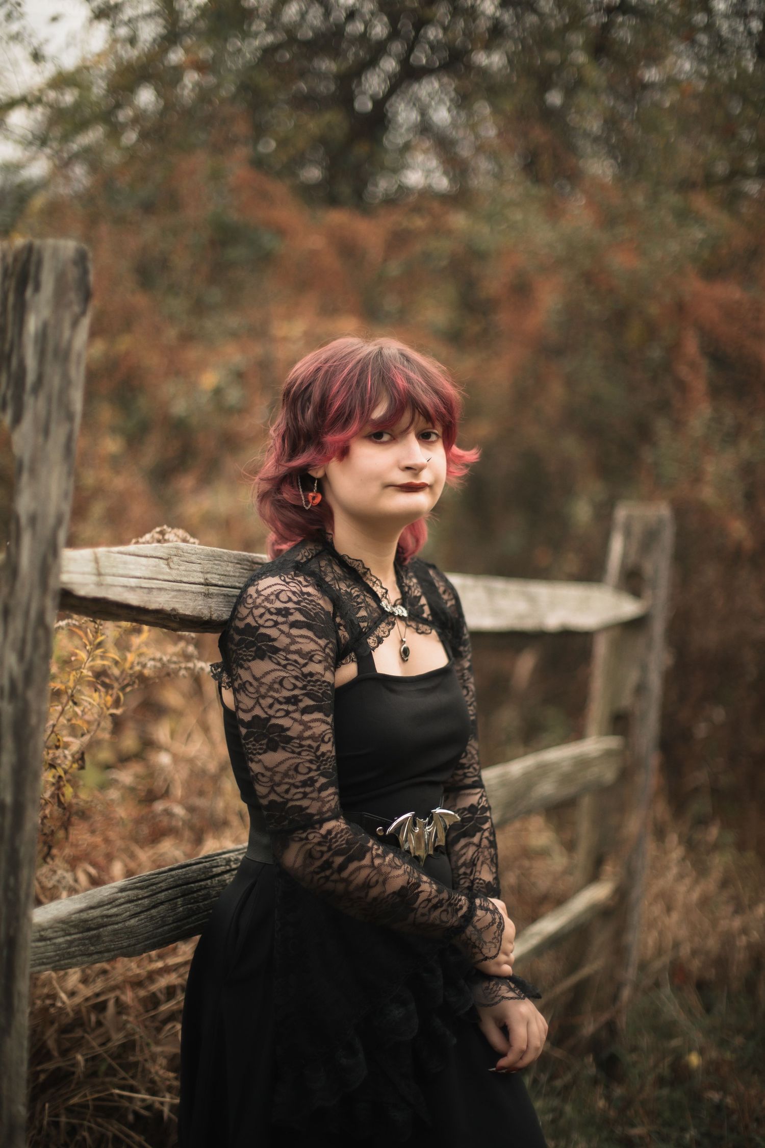 Young woman with red hair in black lace gothic dress leaning against rustic wooden fence in autumn setting.