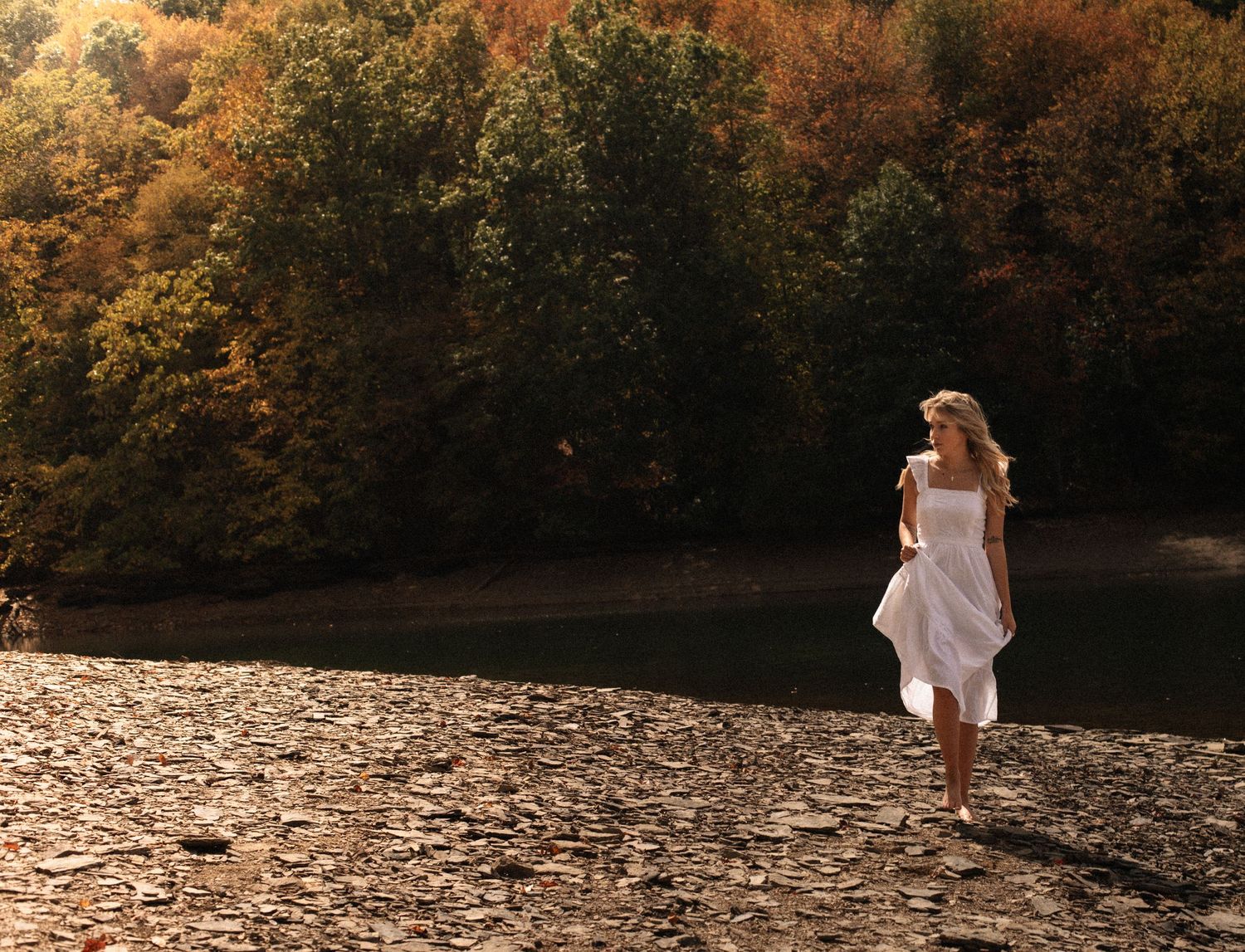 Woman in white dress walking on rocky riverbank surrounded by colorful autumn trees in golden light.