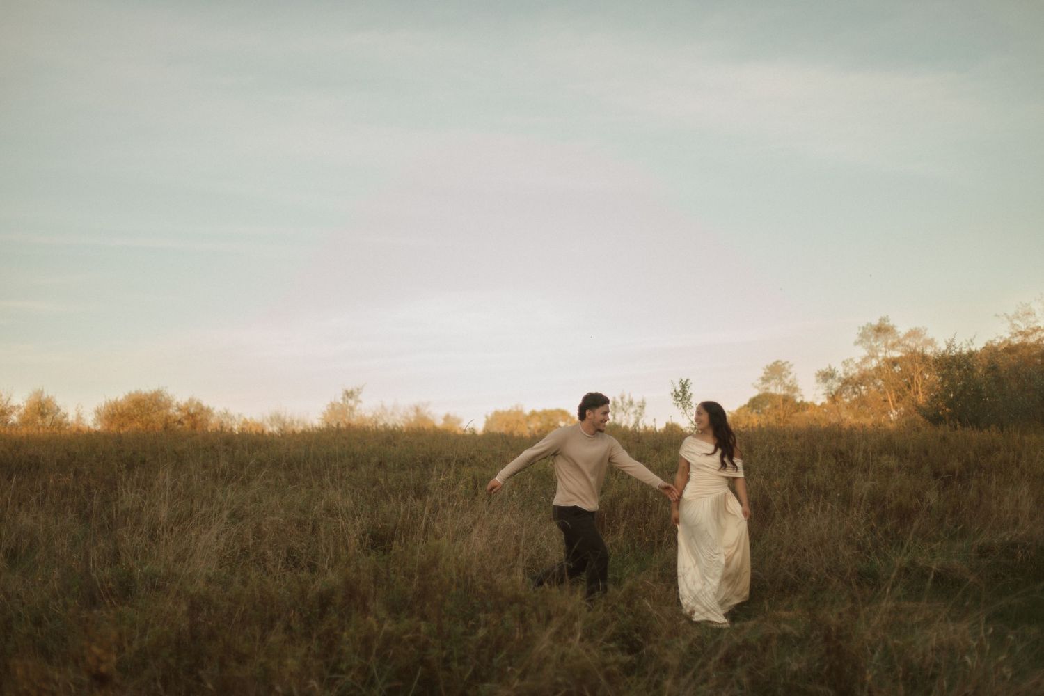 A couple runs hand in hand through a golden field at sunset, creating a romantic silhouette against the warm sky.