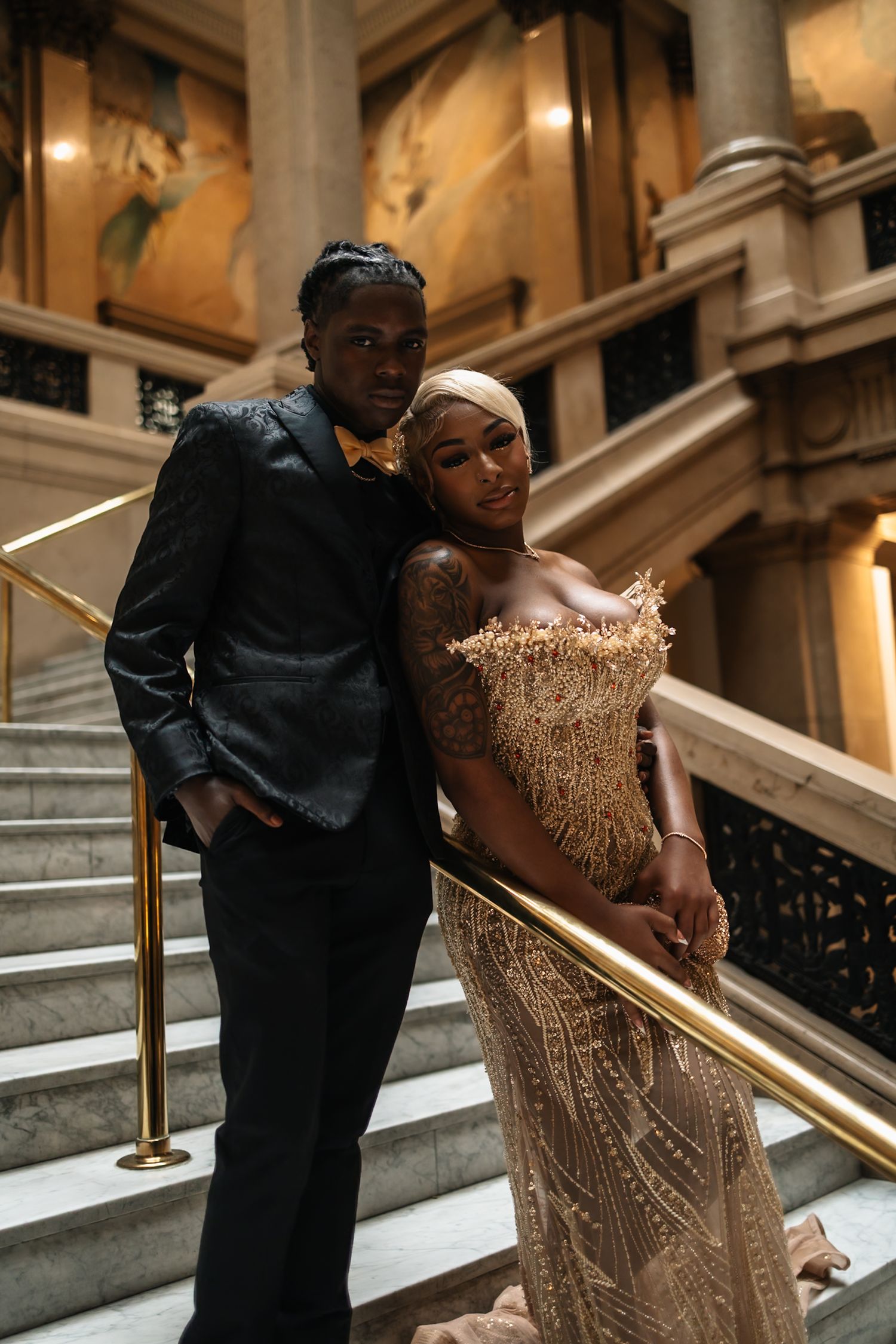 Elegant couple posing on grand staircase, man in black suit, woman in gold beaded gown, formal event.