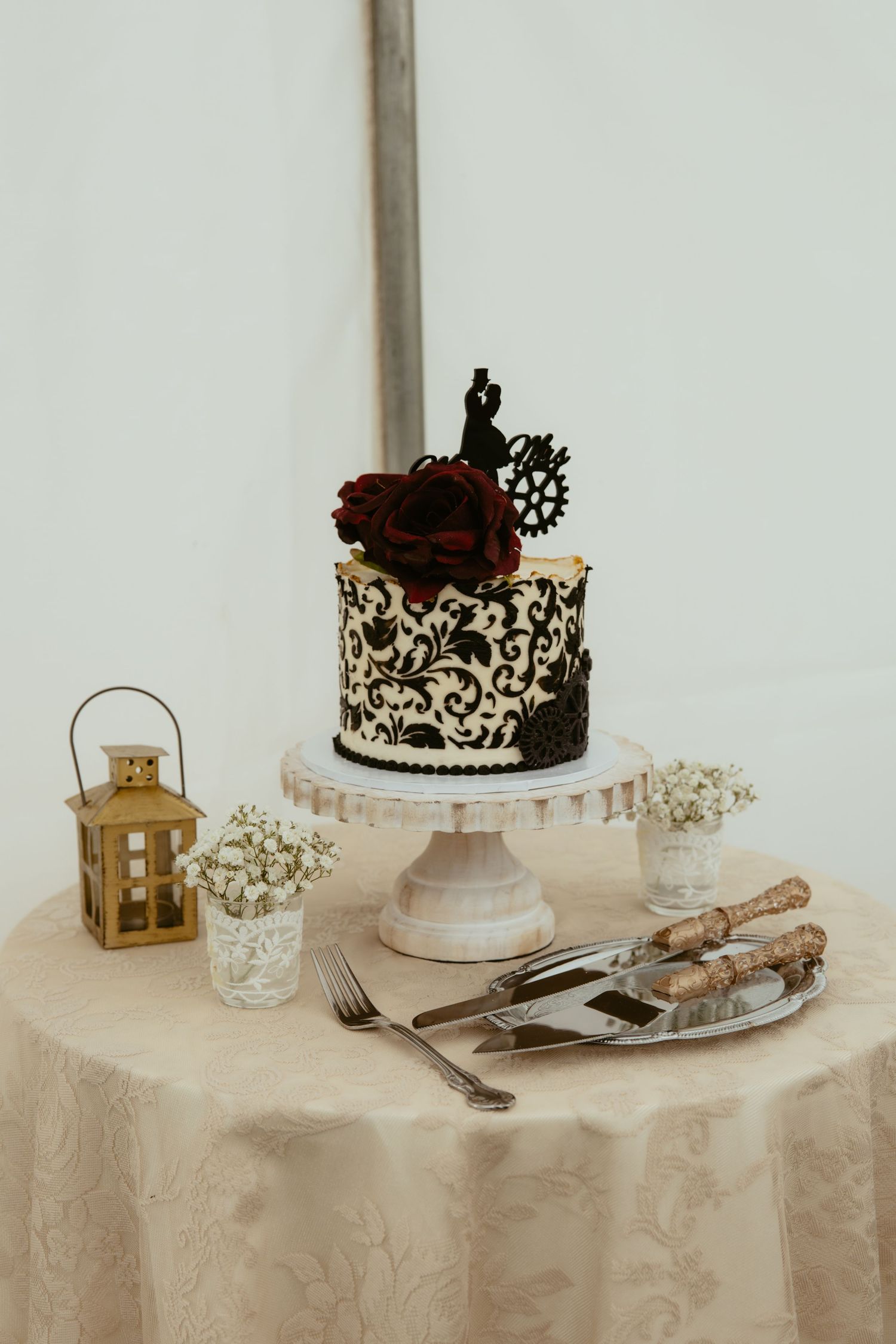 Elegant wedding cake with black damask design, red roses, and couple topper on white stand with lantern and baby's breath.