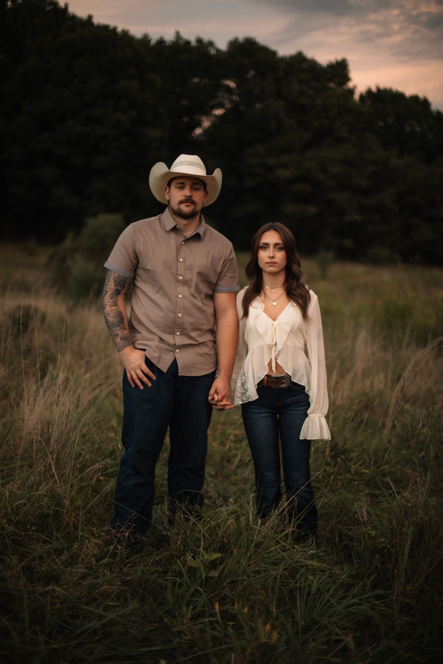 A couple holds hands in a grassy field at dusk, man in cowboy hat and woman in white blouse, with dark trees behind.