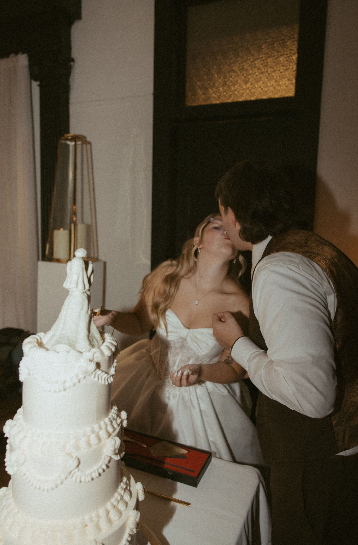 Bride and groom sharing a kiss while cutting their white tiered wedding cake with elegant topper.