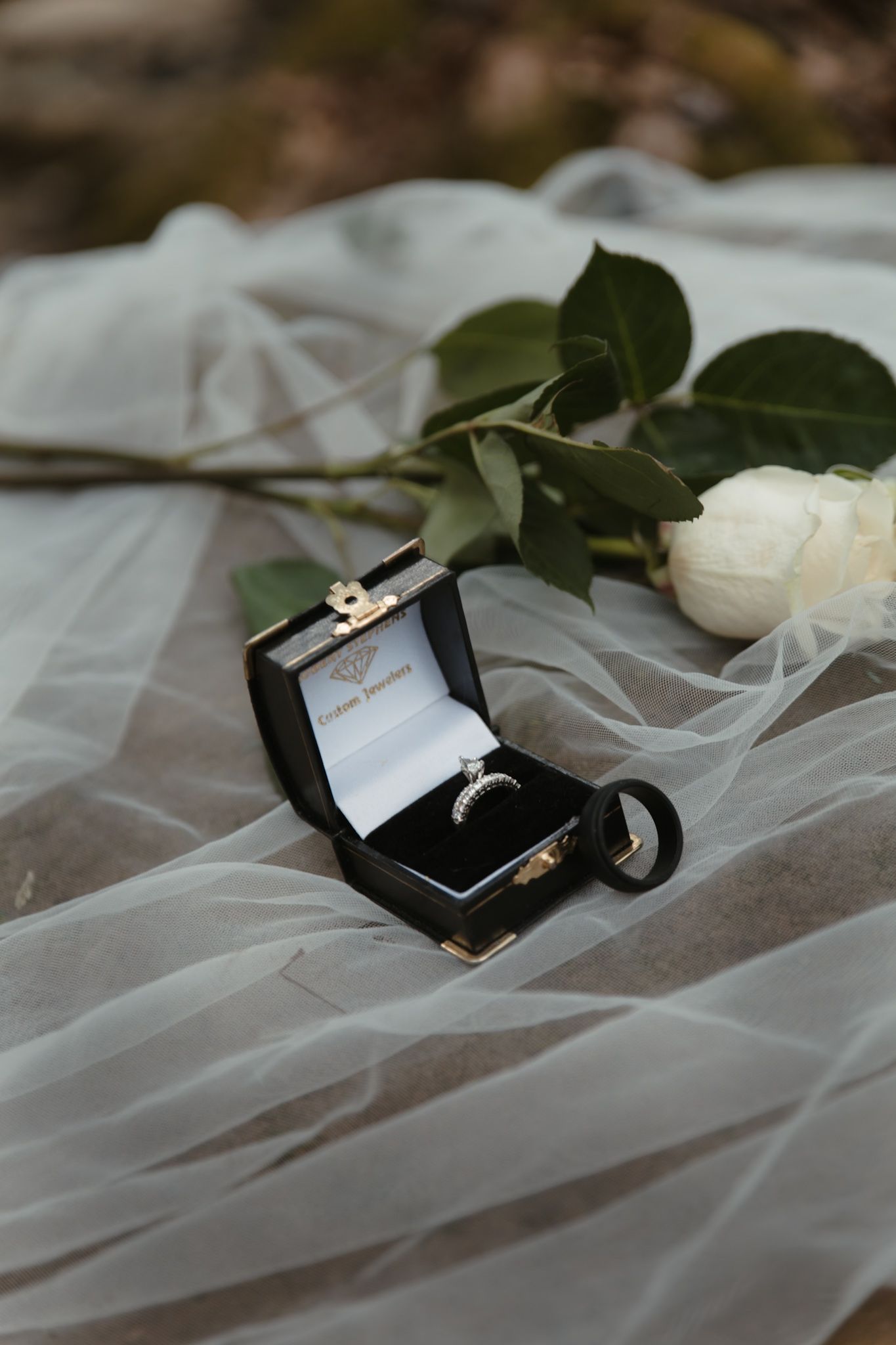 Diamond engagement ring in black box beside white rose on sheer bridal veil.