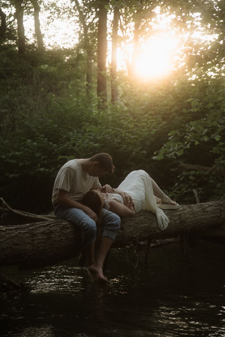 Couple sharing a romantic moment on a fallen log by a river in a sunlit forest at golden hour.
