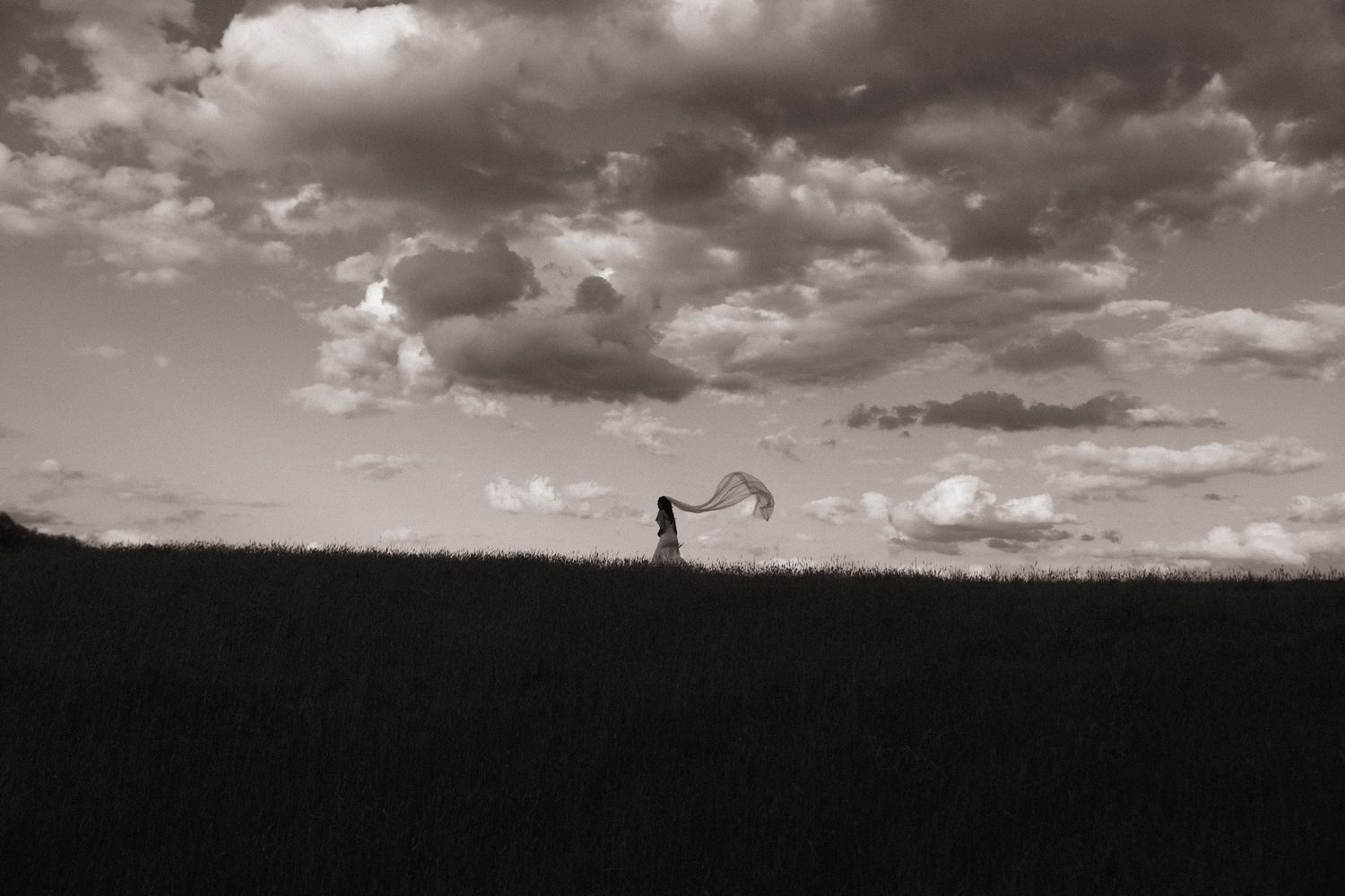 Black and white photo of a lone figure on a hill holding flowing fabric against a dramatic cloudy sky.