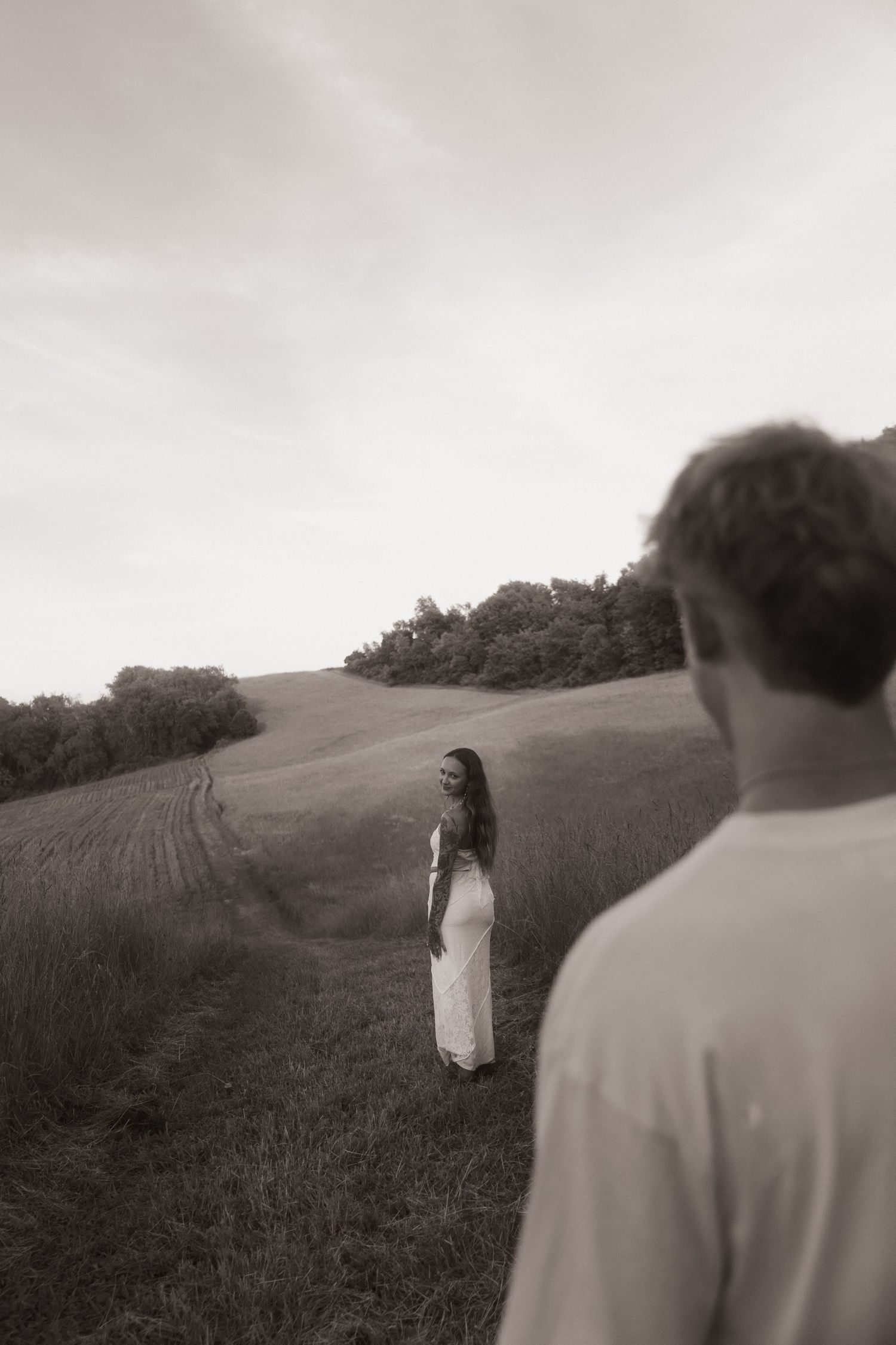 A woman in a white dress stands in a rolling green field while a man watches her from the foreground in sepia tone.