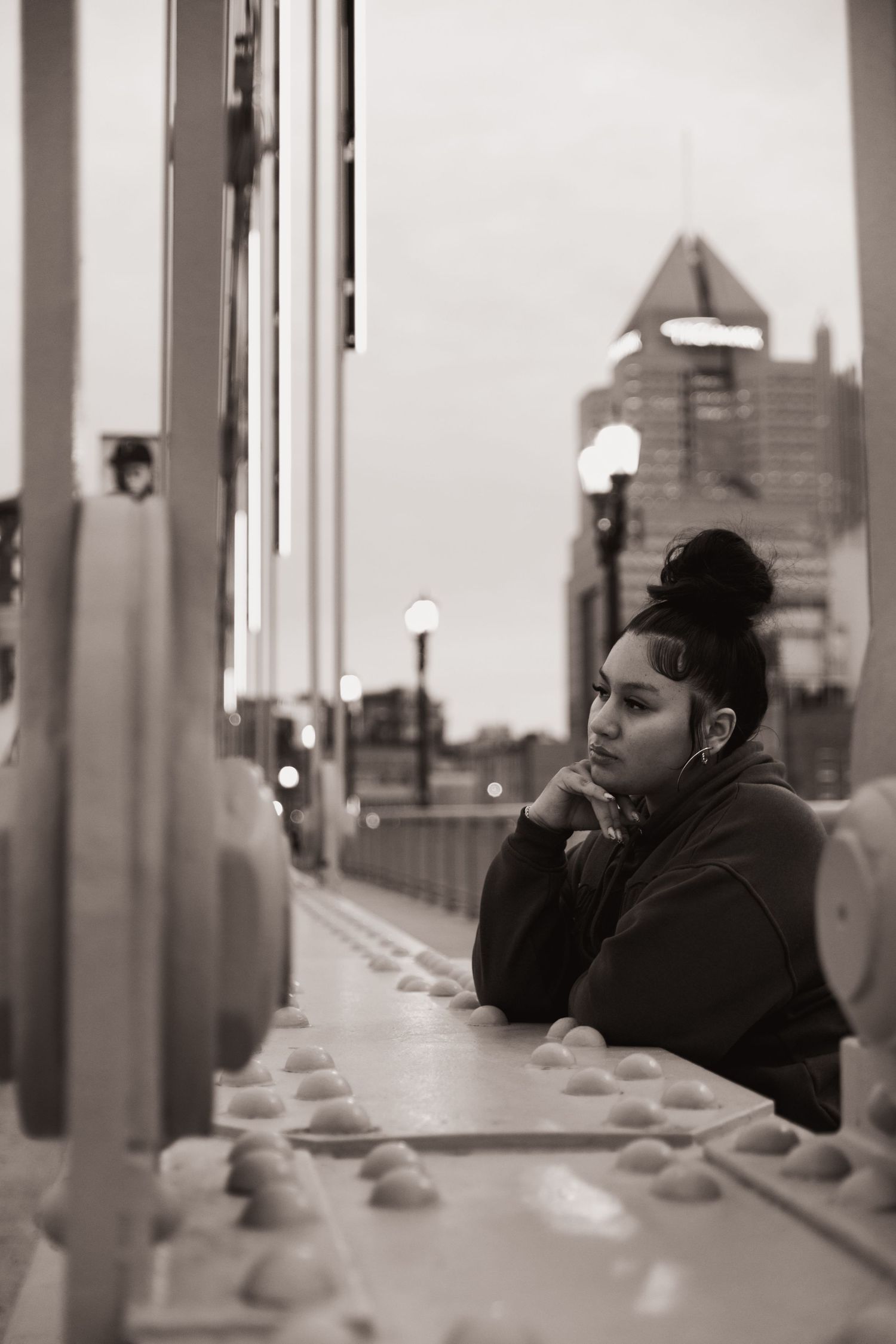 Black and white portrait of a young woman leaning on a bridge railing, city skyline visible in background.