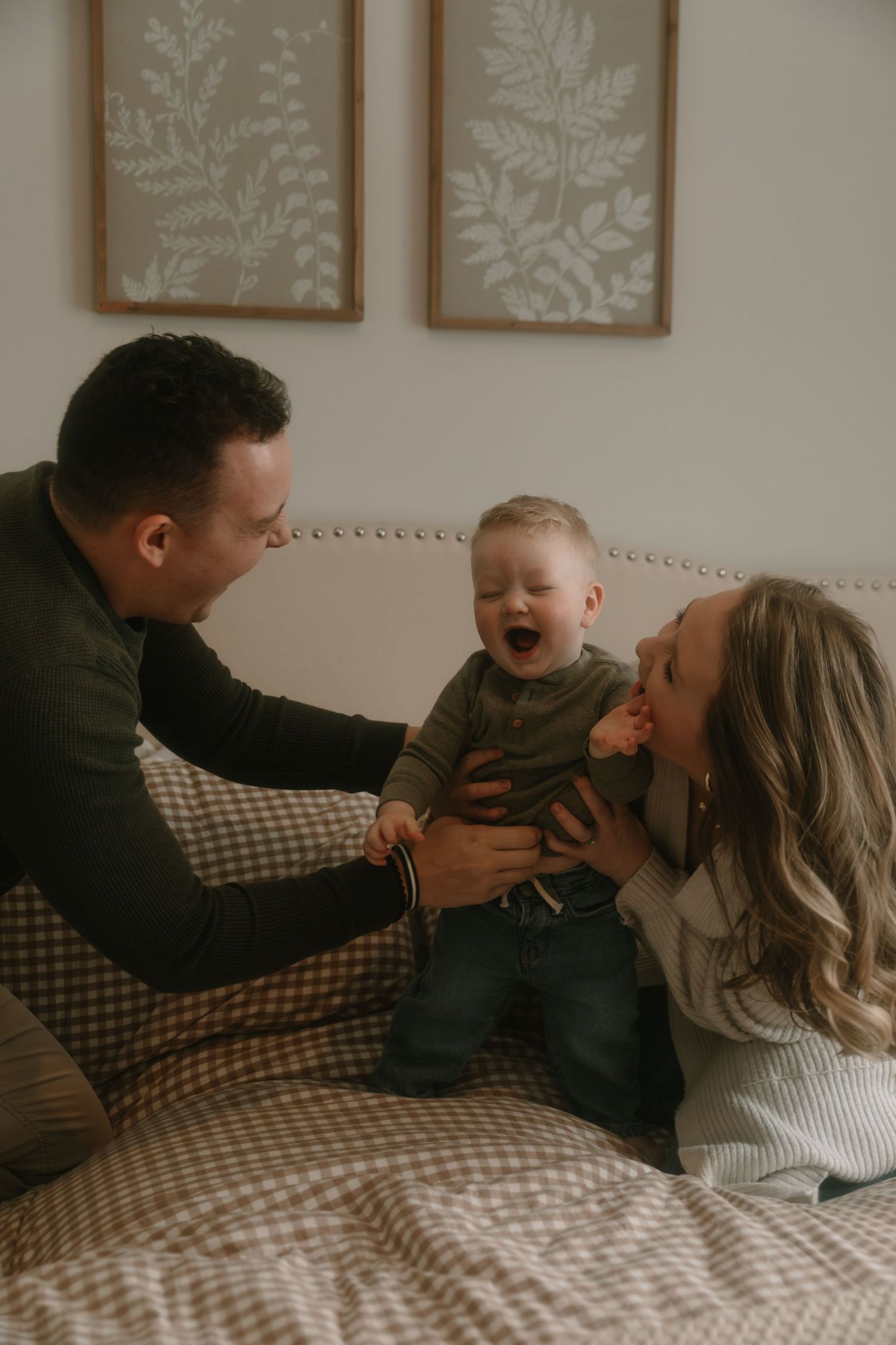 Happy family with laughing baby boy sitting on bed with botanical wall art in cozy neutral bedroom setting.