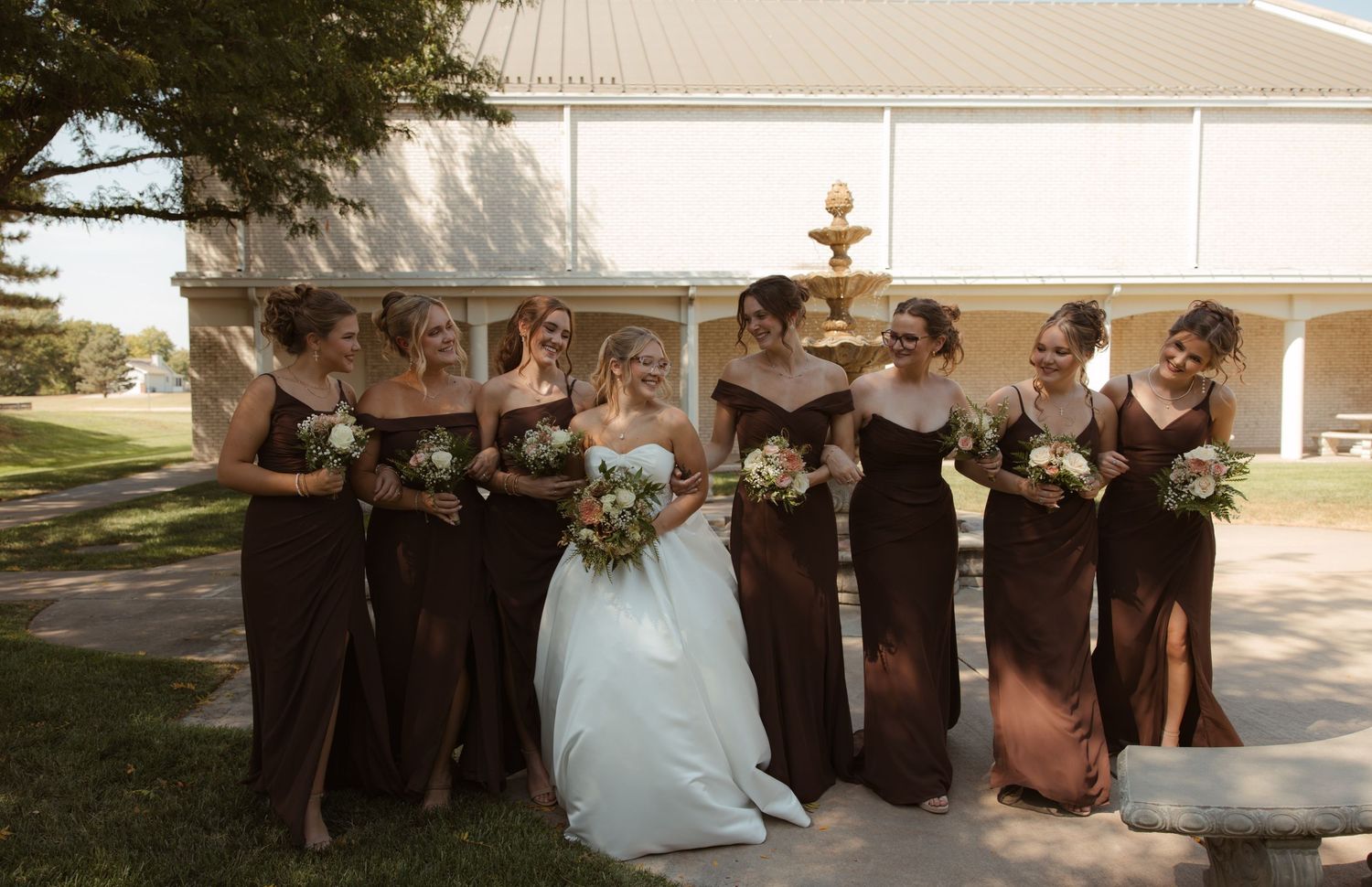 Bride in white gown with bridesmaids in brown dresses holding bouquets outside a white building on a sunny day.