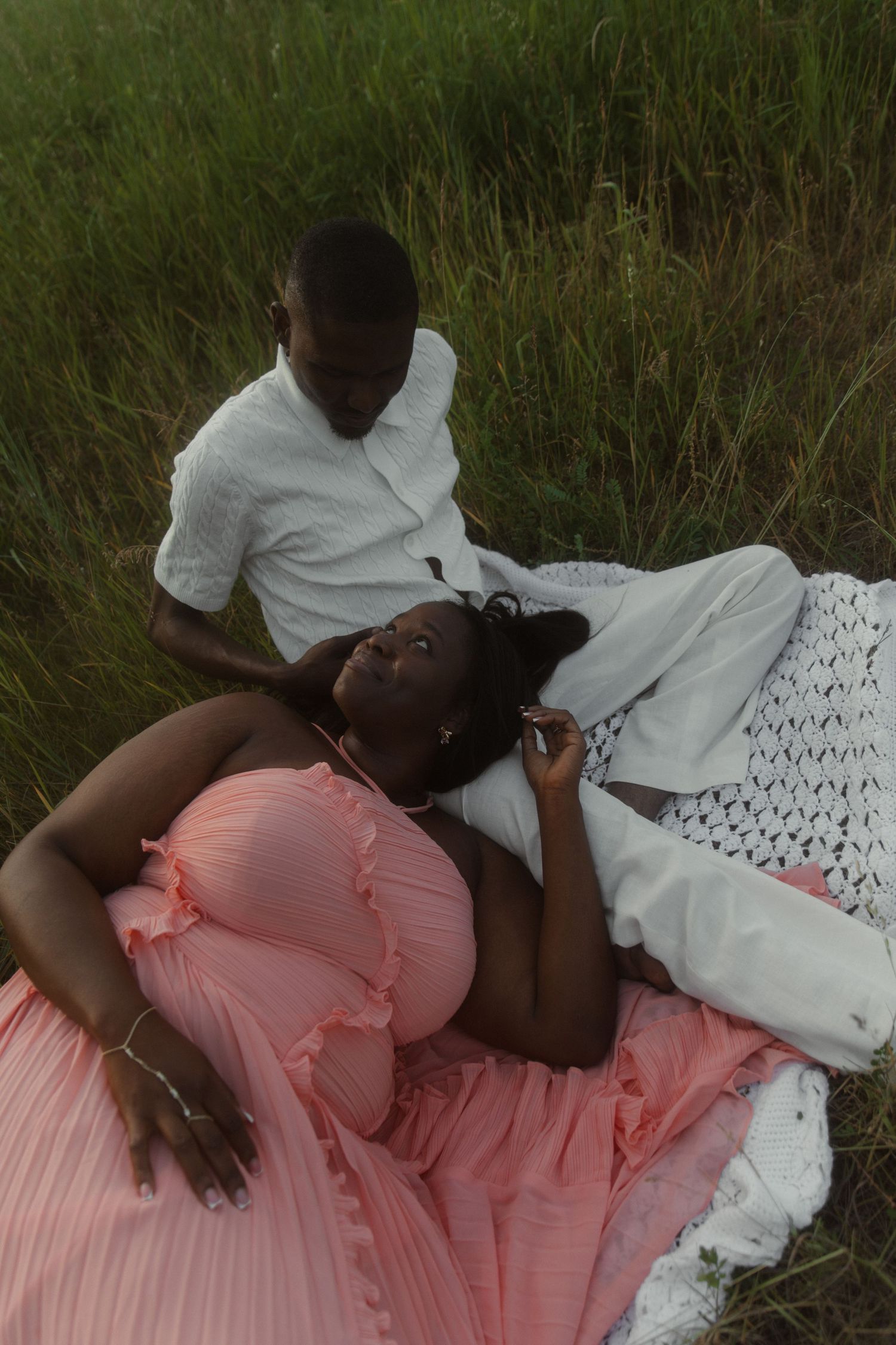 A pregnant woman in a pink dress lies in a field while her partner in white sits beside her.