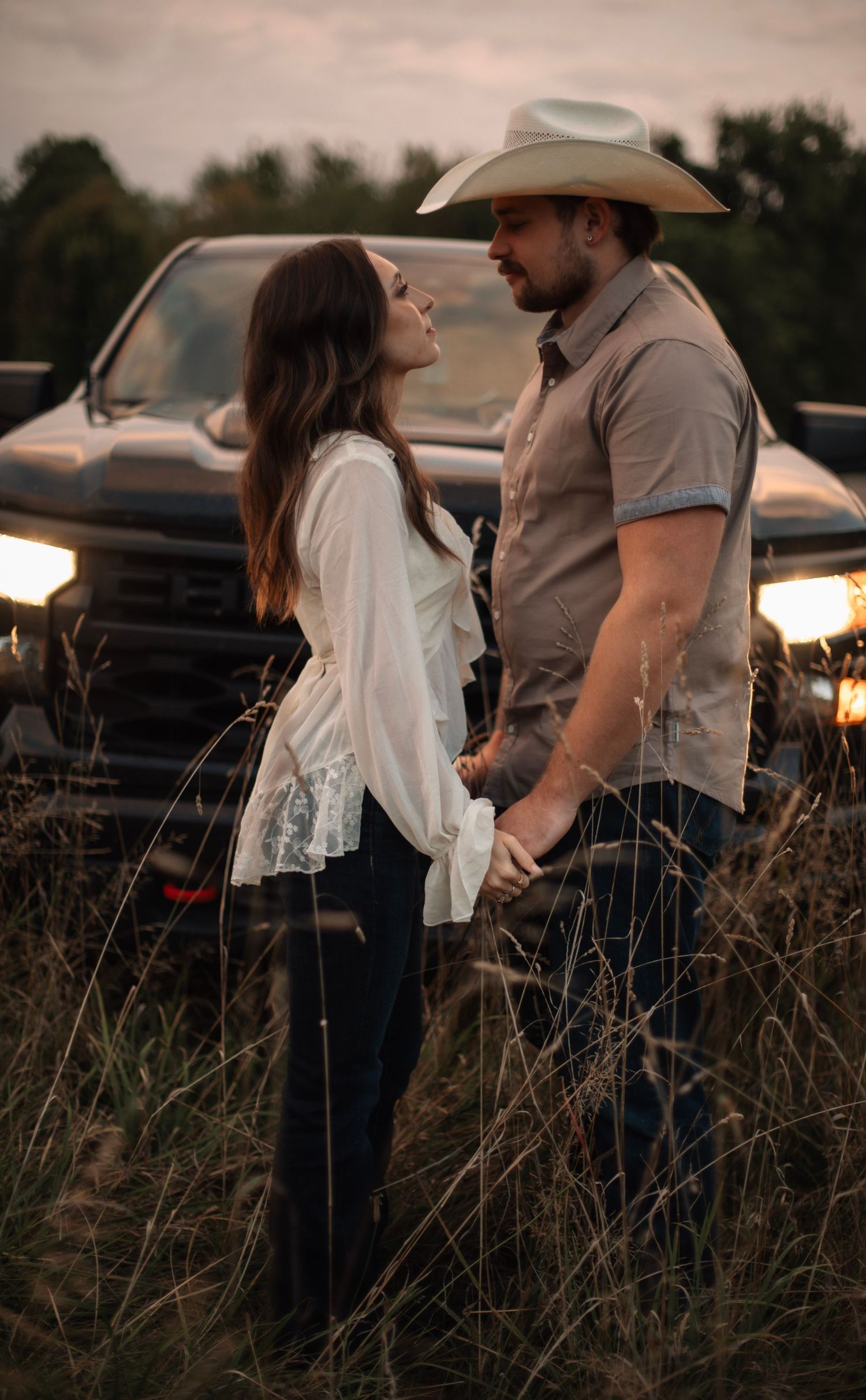 A couple shares an intimate moment in a field at sunset with a cowboy hat and vintage car in view.