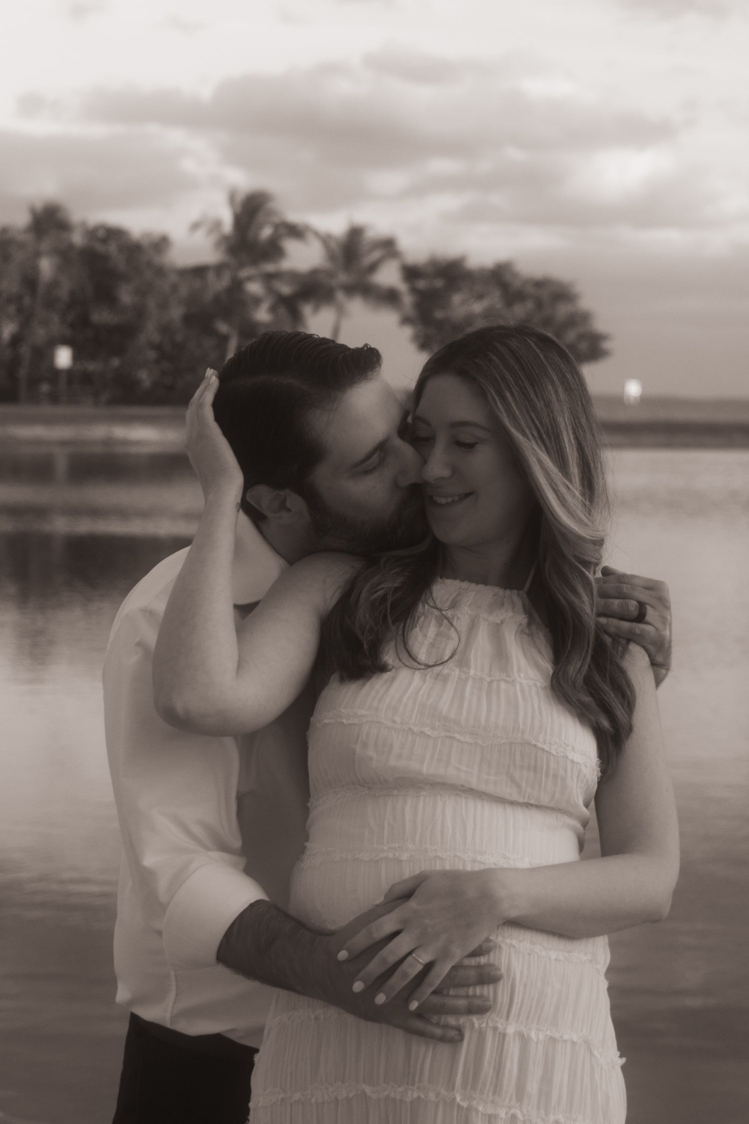 A romantic black and white photo series showing a couple embracing by a serene waterfront at sunset.