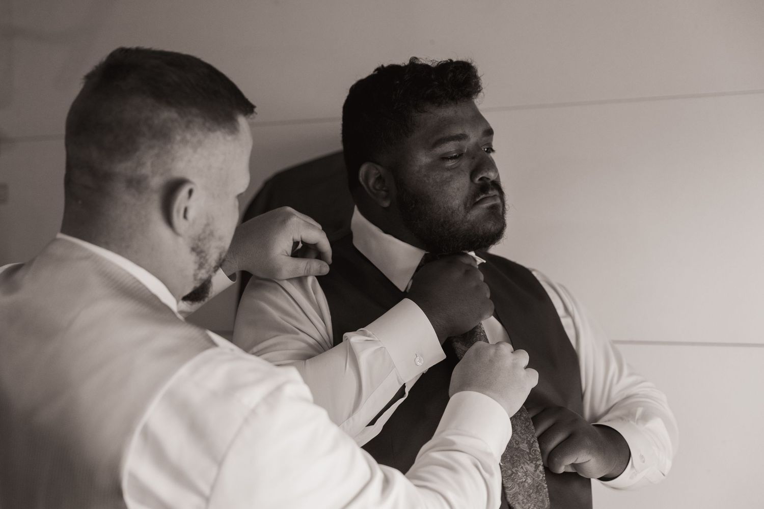 Black and white photo of a man helping another man adjust his tie, both dressed in formal wedding attire.