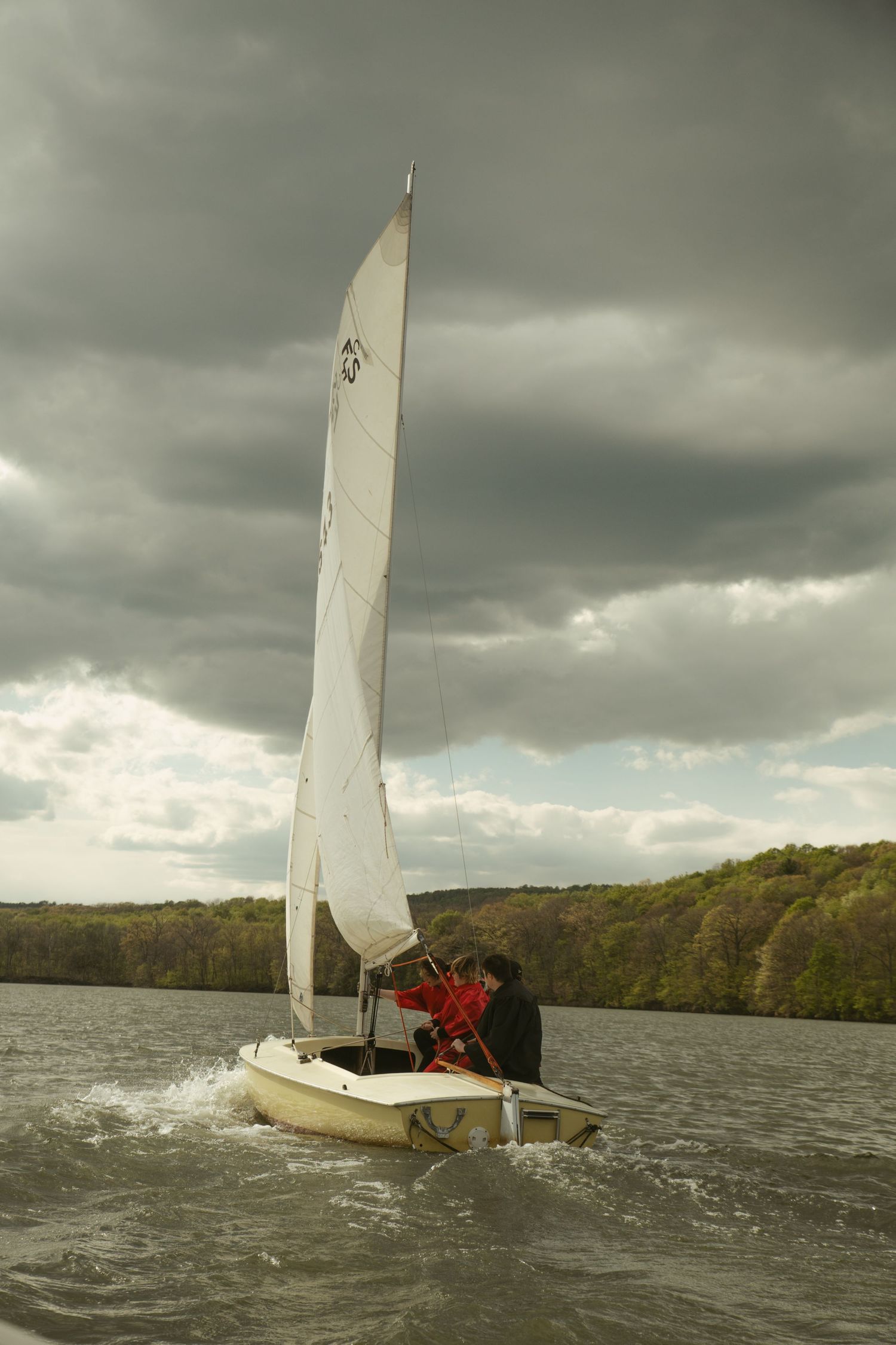 Two sailors in red jackets navigate a white sailboat on choppy waters under dramatic storm clouds.
