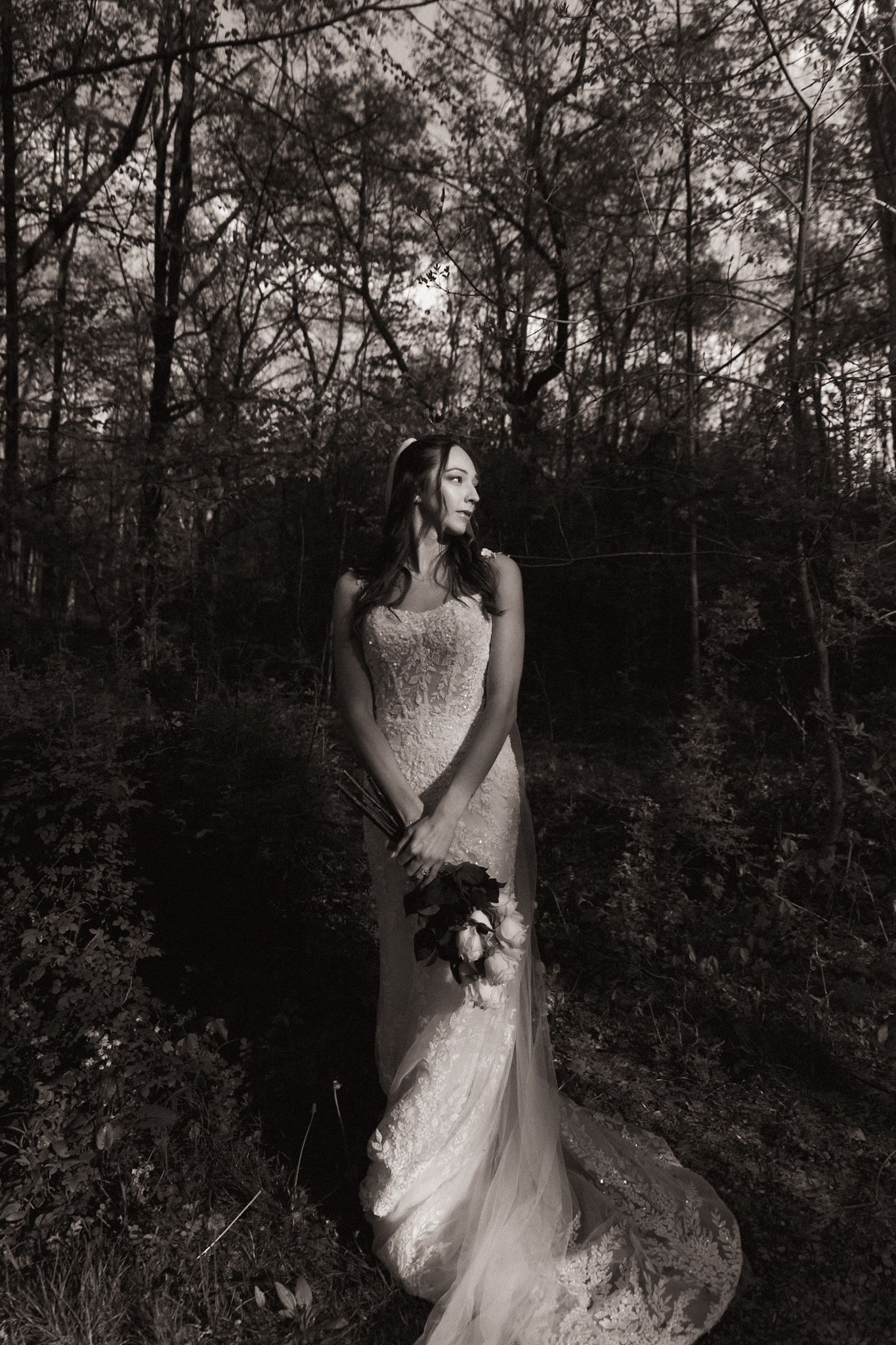 Black and white photo of a bride in a lace gown holding flowers in a dark forest setting.
