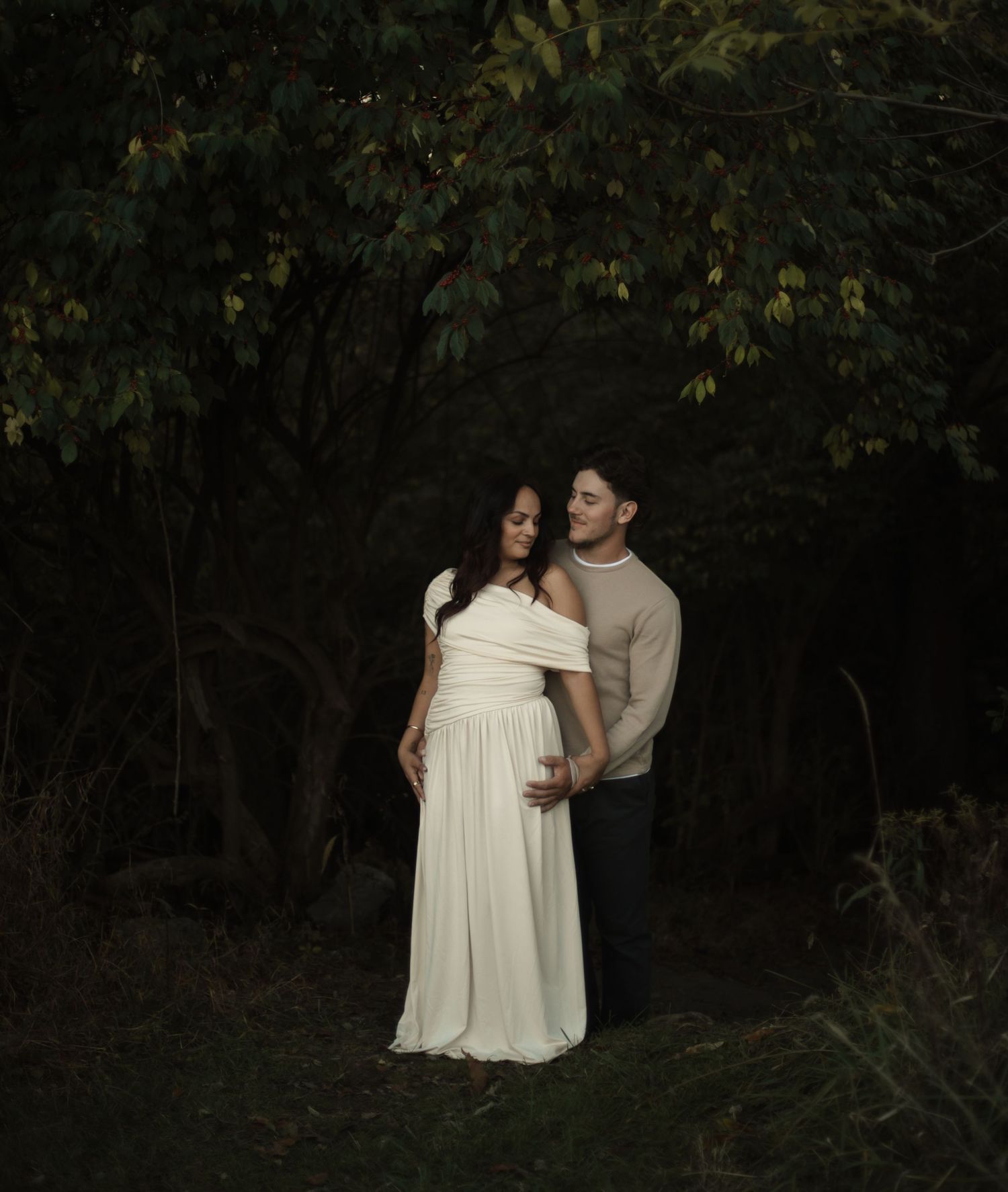 Couple embracing in a dark forest, woman in white off-shoulder gown, man in beige sweater, maternity photo shoot.