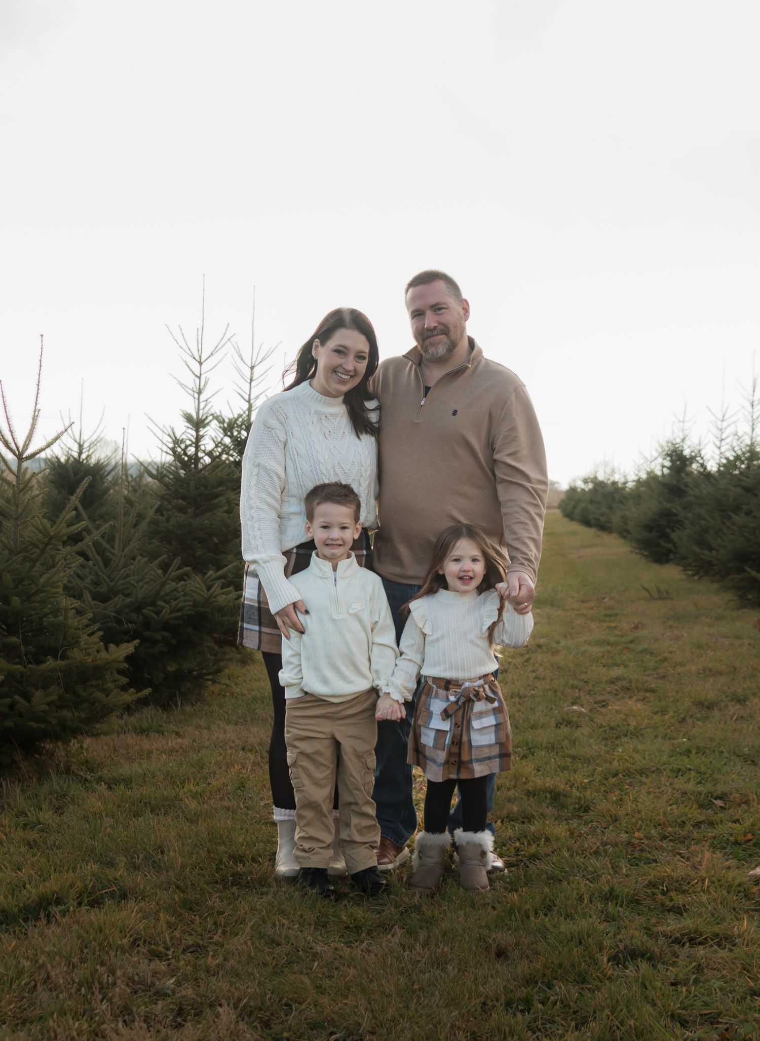 A family in matching tan and white outfits poses together at a Christmas tree farm on a cloudy day.