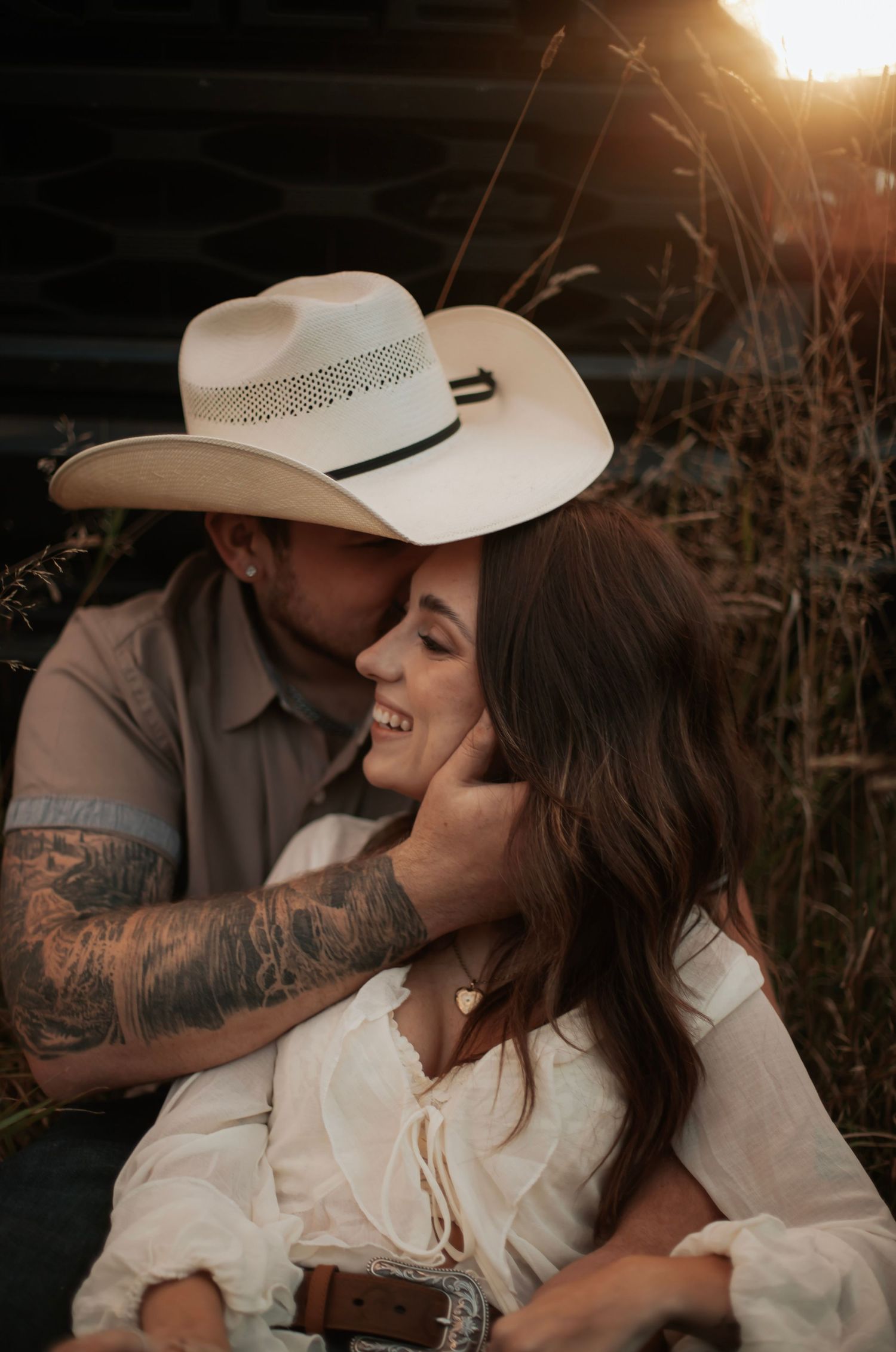 Couple sharing a romantic golden hour moment, man in cowboy hat with tattoos kissing woman in white blouse.
