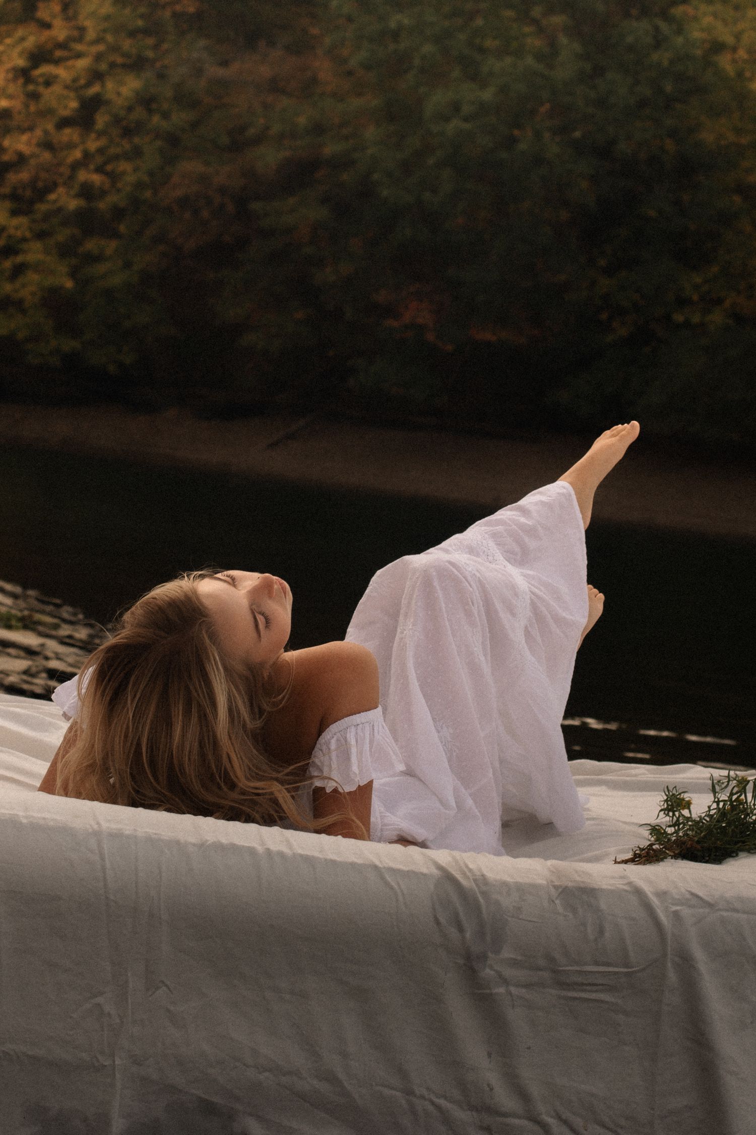 Woman in white dress lying on white fabric outdoors near a river surrounded by autumn trees.