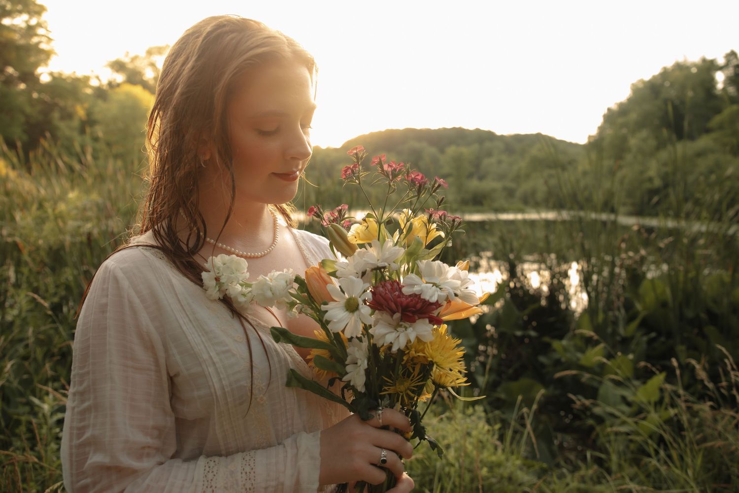 Woman holding a bouquet of wildflowers in golden sunset light near a serene pond surrounded by lush greenery.