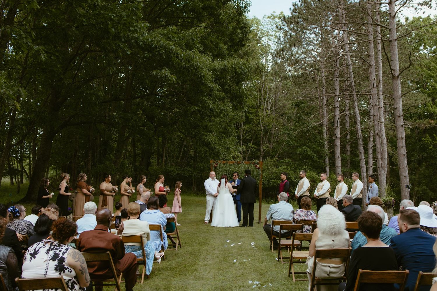 Outdoor wedding ceremony on a lush green lawn surrounded by tall trees, with guests seated in wooden chairs facing the couple.