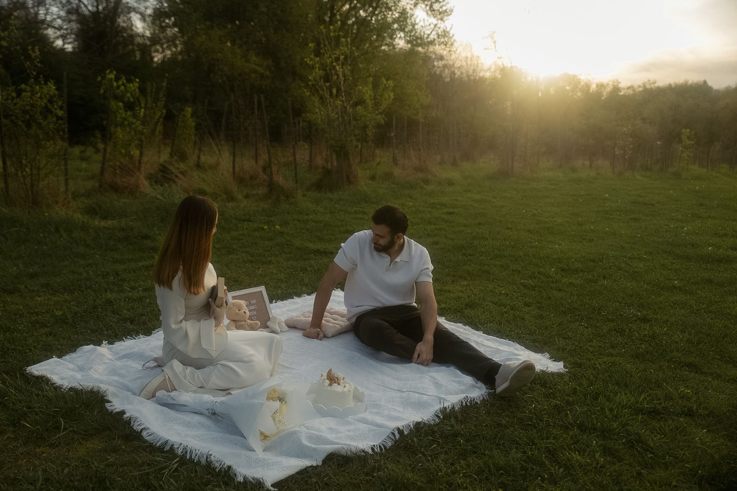 Couple sitting on white blanket having a romantic picnic in green meadow at golden sunset hour.