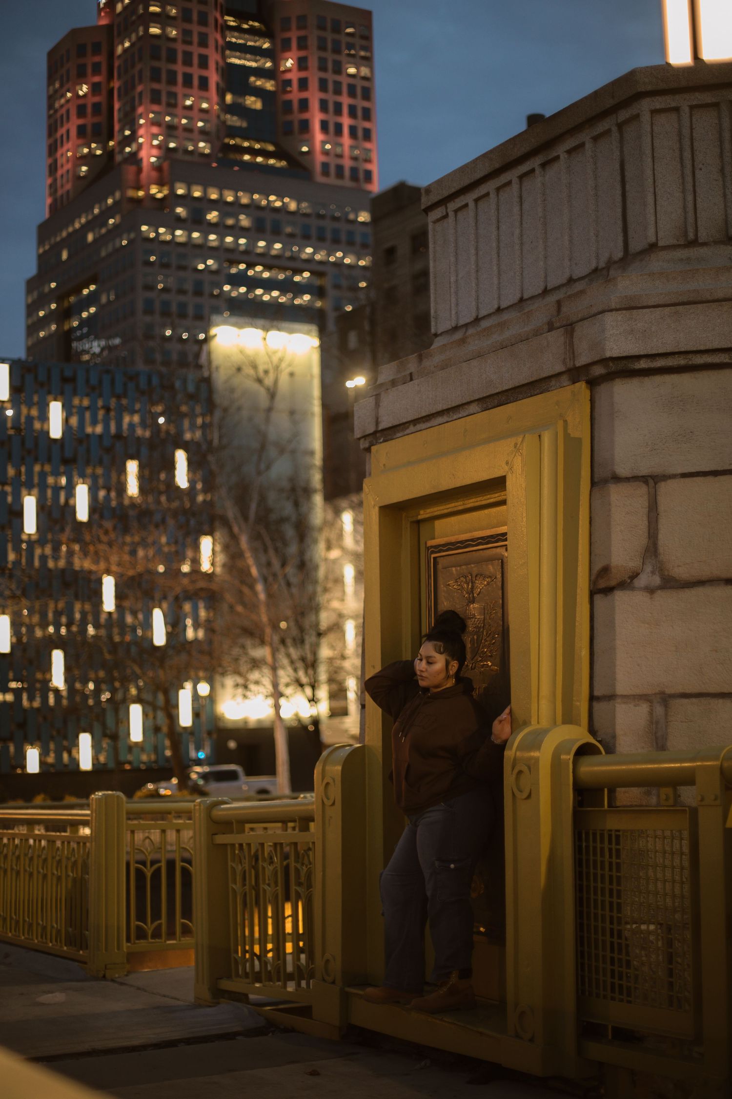 Woman poses by ornate golden door on urban bridge at dusk with illuminated skyscrapers in background.