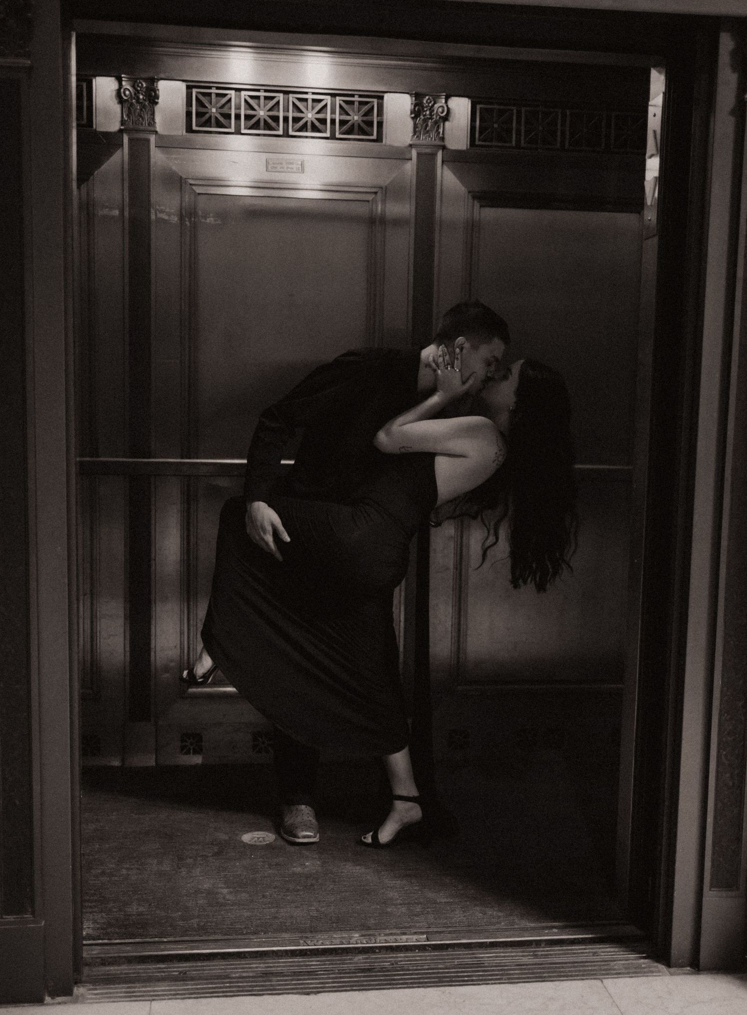 Couple sharing a romantic kiss in an elegant elevator doorway, captured in black and white photography.