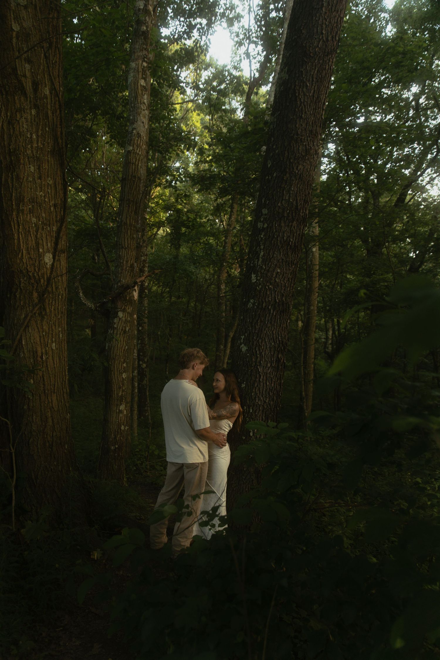 Romantic couple embracing in a lush green forest, standing among tall trees in soft natural light.