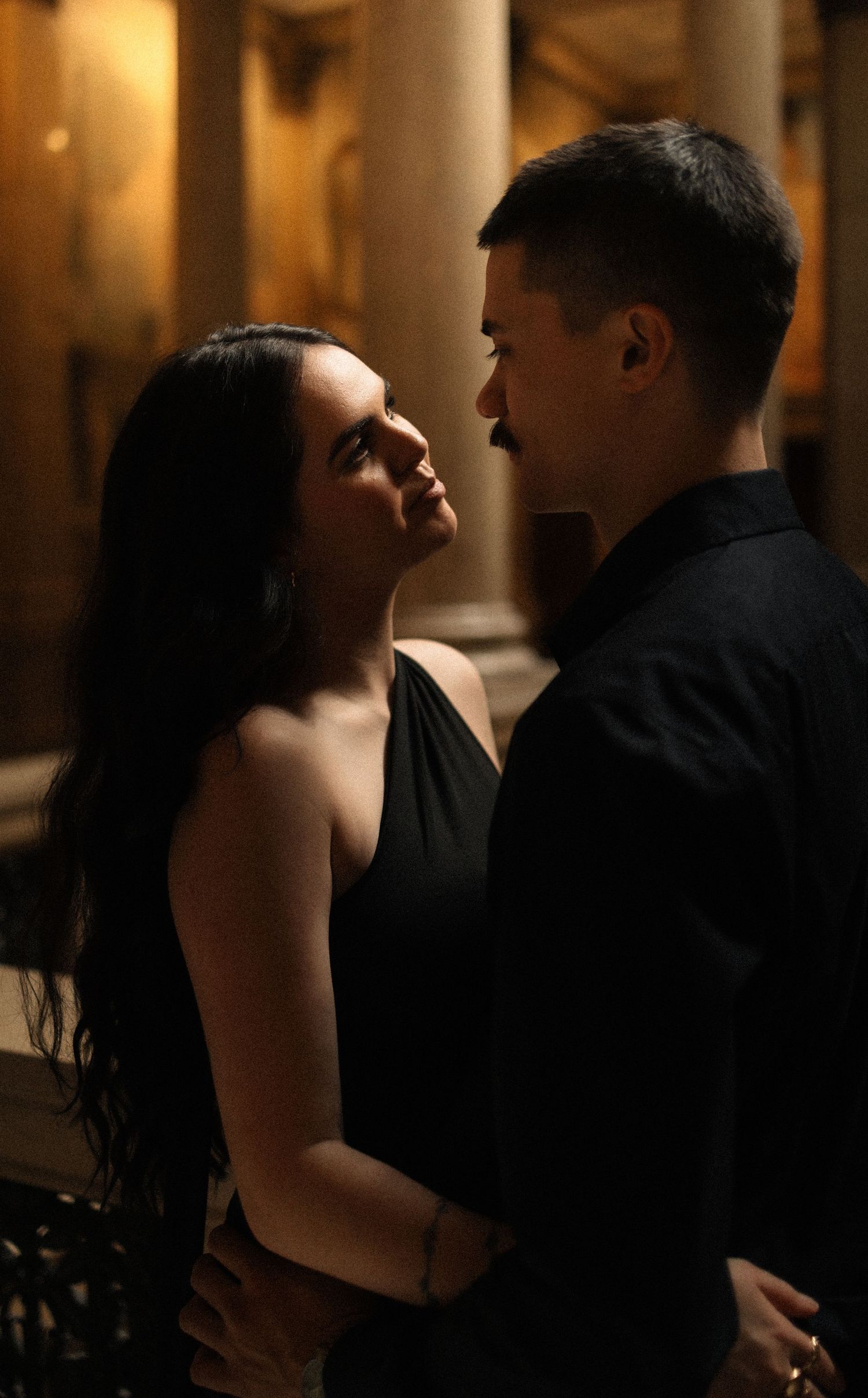 A romantic couple in black attire share an intimate gaze in a dimly lit classical interior with stone columns.