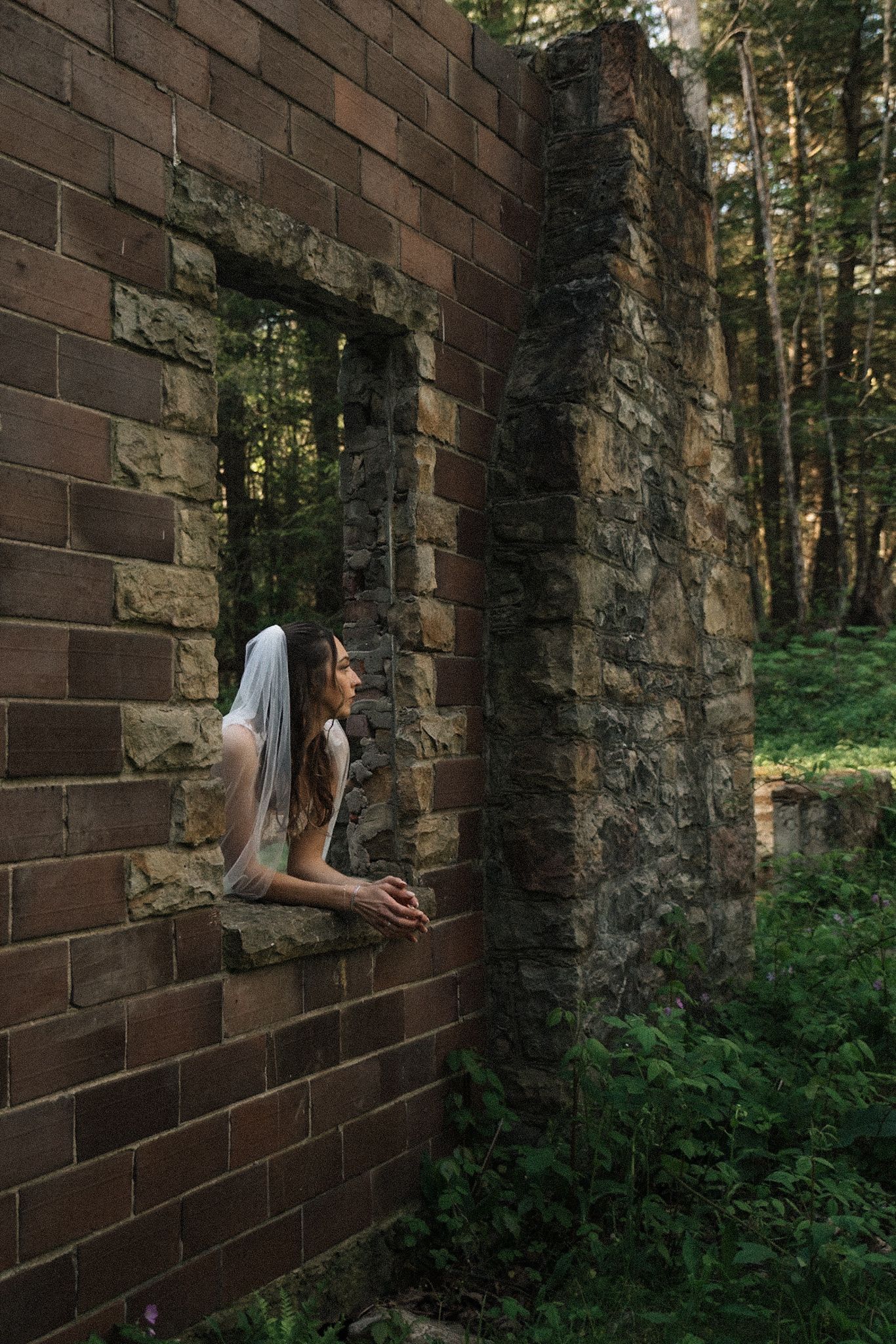 Bride in white veil gazes from window of rustic brick ruins surrounded by lush forest.