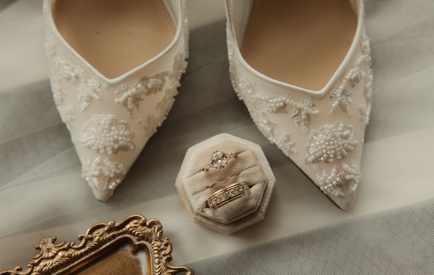 Elegant white lace bridal heels with pearl embellishments beside a velvet ring box with diamond wedding rings.