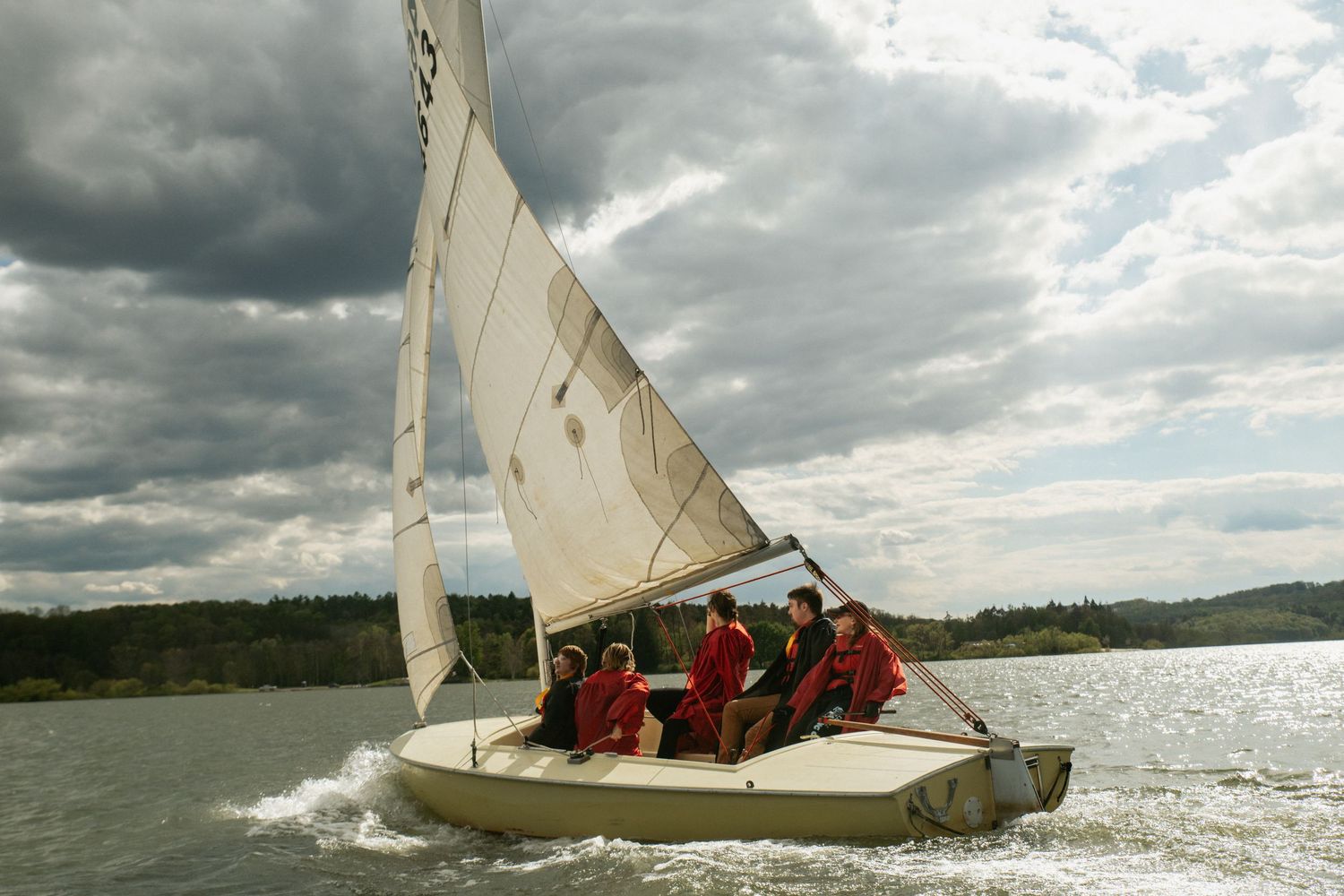 Group of people in red jackets sailing on a white sailboat across a calm lake under dramatic cloudy skies.