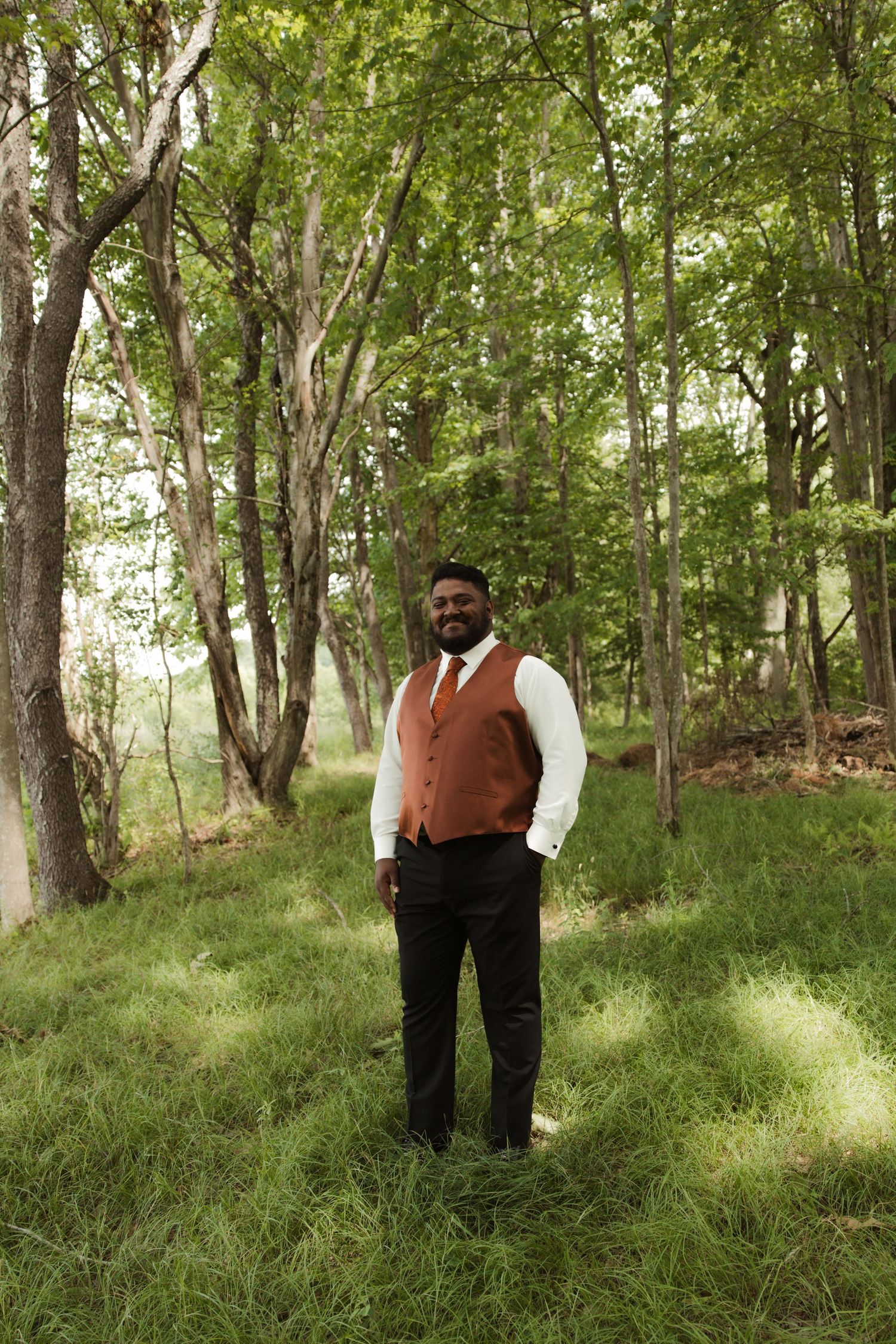A smiling groom in a rust vest and white shirt stands in a lush green forest clearing on his wedding day.