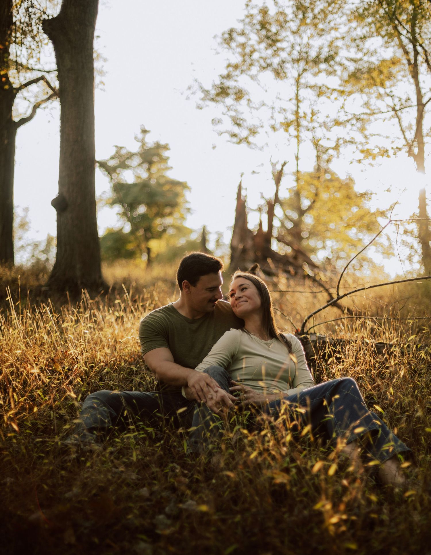 A couple shares a tender moment while laying in tall grass during golden hour in a wooded area.