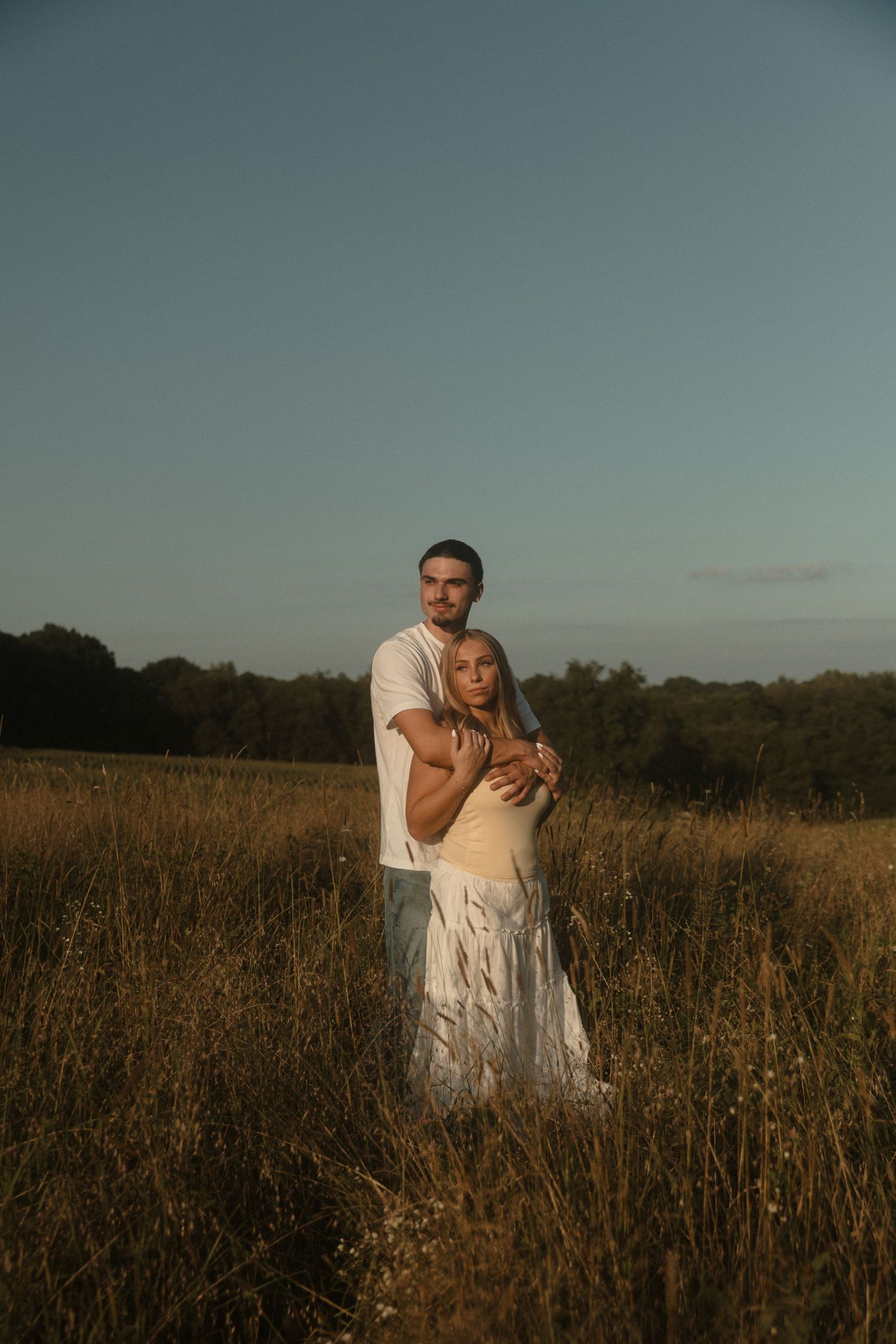 Couple embracing in golden field at dusk, man in white shirt hugging woman in white skirt, trees in background.